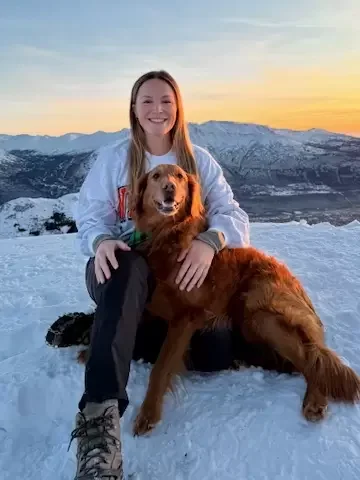 A young woman with long hair smiling and sitting on snow with a golden retriever dog during sunset in a snowy mountain landscape.