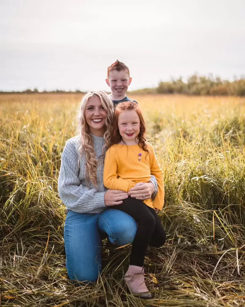 A woman kneeling in a field with two children, a girl in a yellow sweater and a boy standing behind them, smiling at the camera.