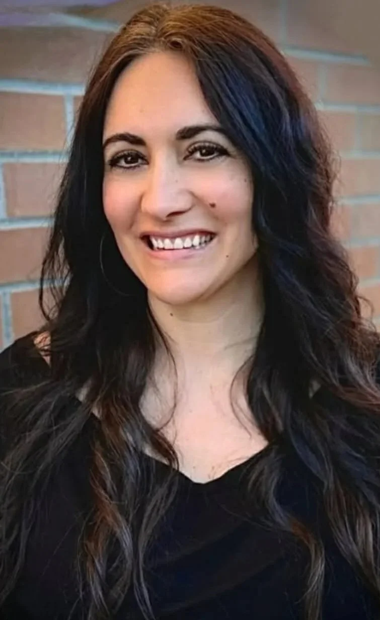 A young woman with long dark wavy hair and light skin smiling in front of a brick wall.