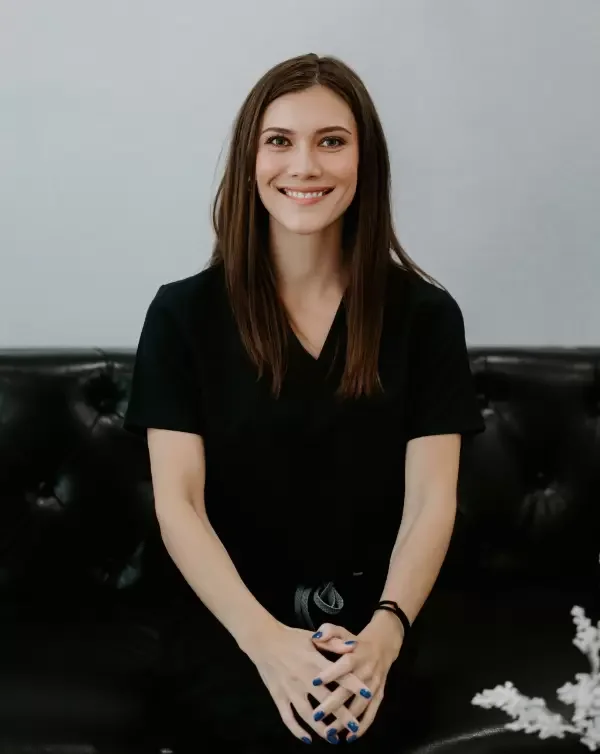 Young woman with long brown hair smiling, sitting on a black leather couch, wearing a black t-shirt and blue nail polish, with a light gray wall in the background.