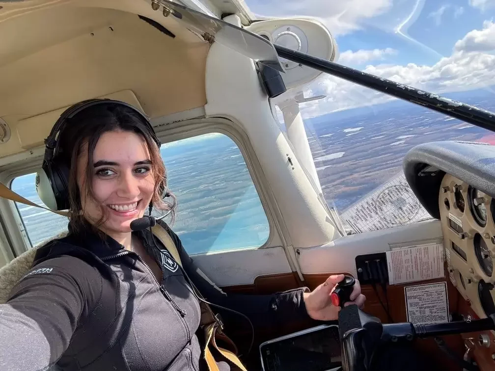 A smiling female pilot sitting in the cockpit of a small airplane, wearing a black jacket and a headset, with an outside view of the sky and landscape through the windows.