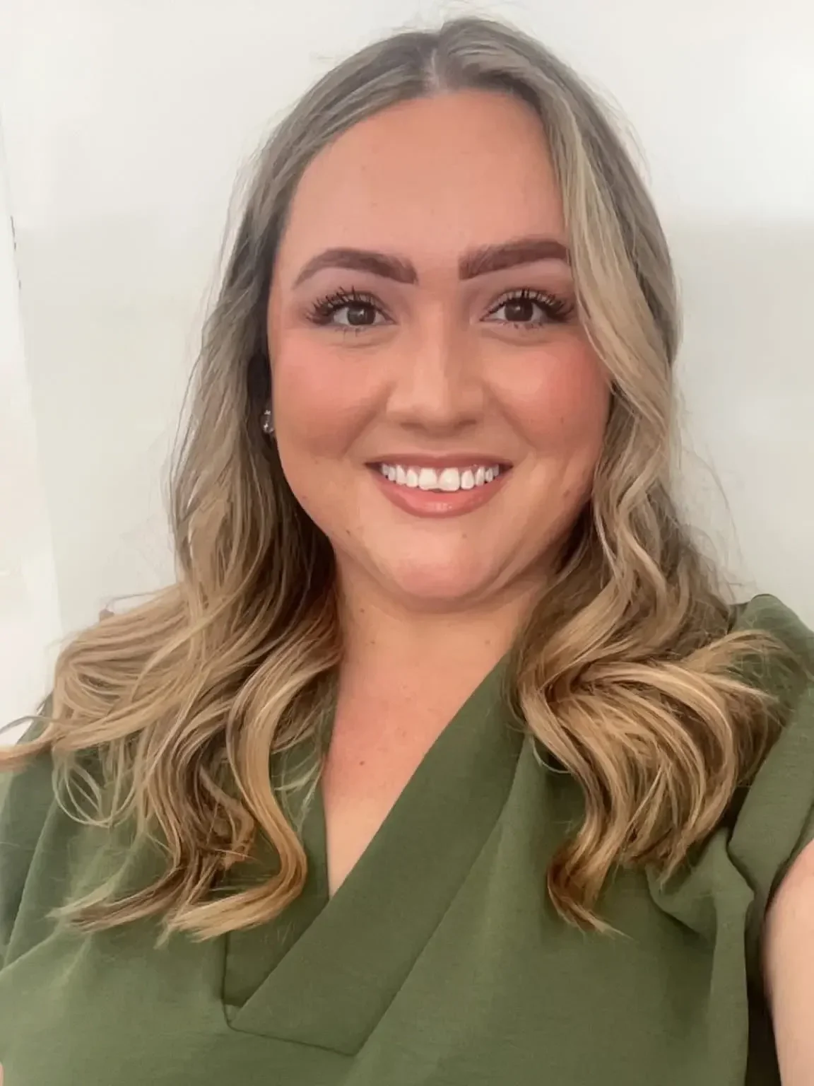 Close-up of a smiling woman with wavy blonde hair wearing a green top, standing against a white background.