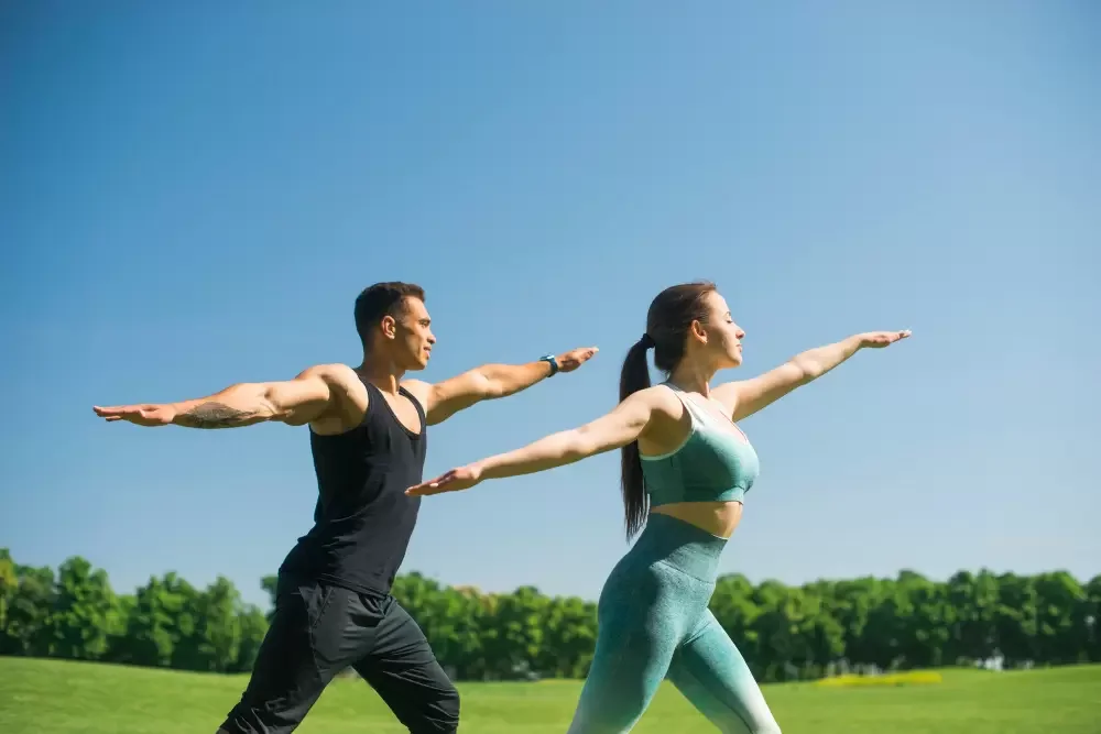 A man and woman doing yoga outdoors in a park, stretching with arms extended forward, under a clear blue sky.