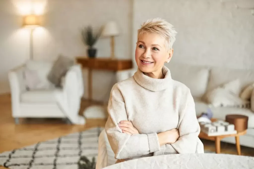 A woman with short blonde hair smiling and crossing her arms, sitting in a cozy, well-lit living room with neutral-colored furniture and decor.
