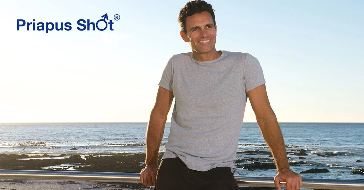 A smiling man in a gray T-shirt leaning on a railing at the beach during sunset, with the ocean in the background and the logo 'Priapus Shot' in the top left corner.