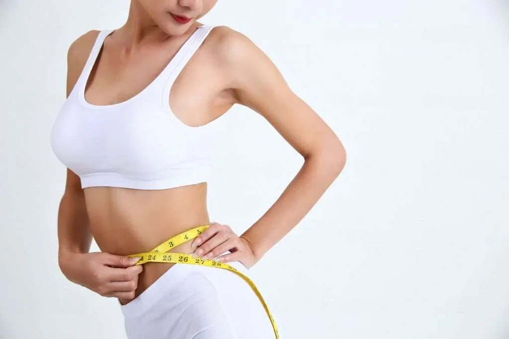 A woman in a white sports bra and shorts measuring her waist with a yellow tape measure.