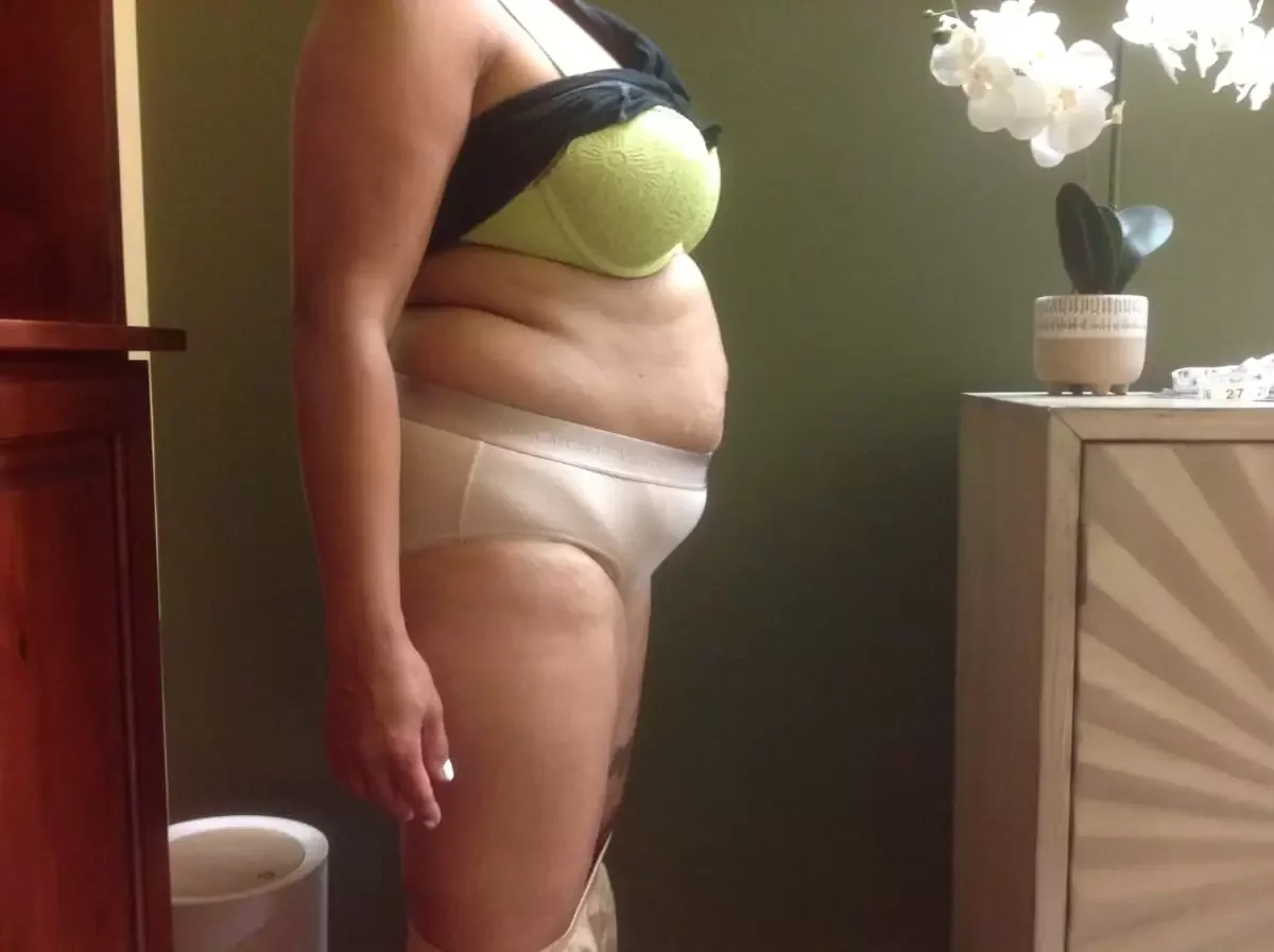Side view of a woman standing indoors, wearing a green bra and beige underwear, with flowers on a table nearby.