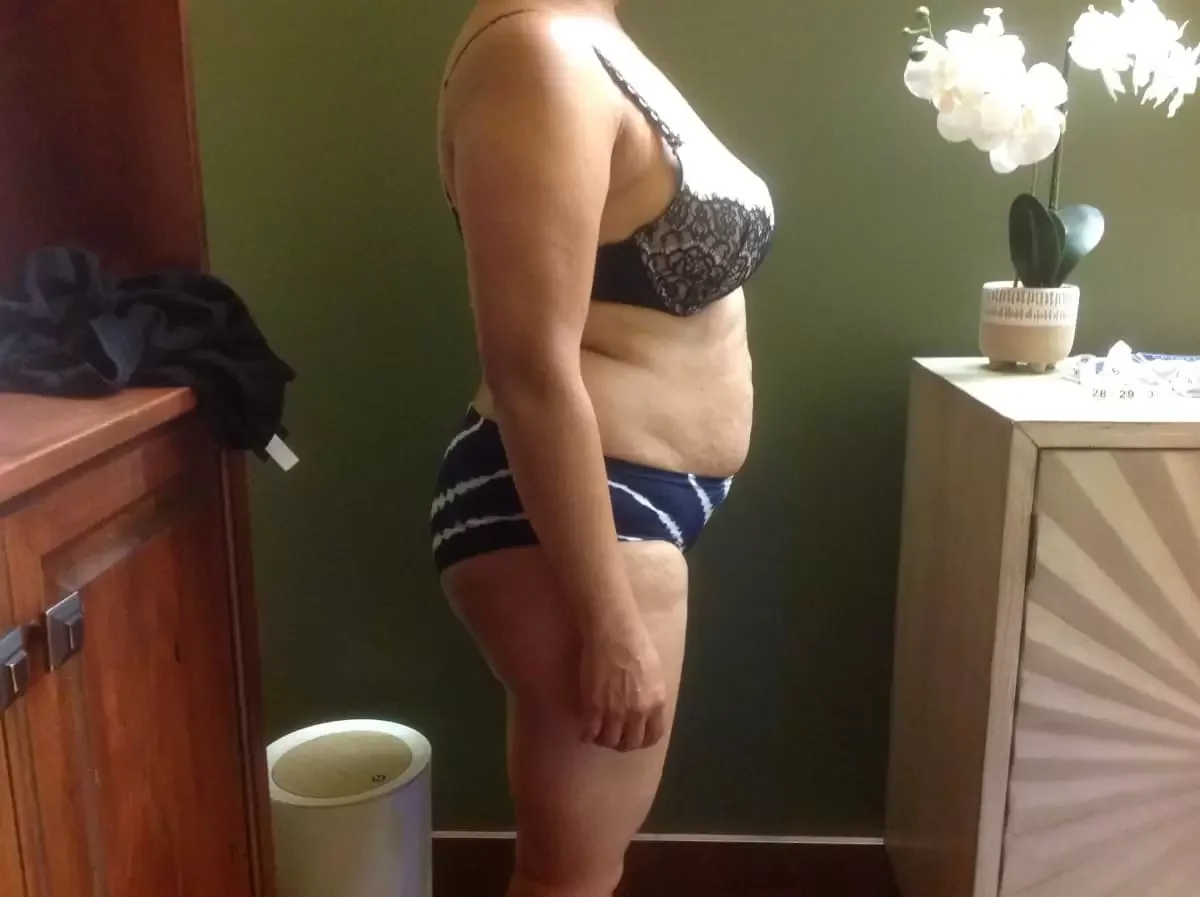 Profile of a woman standing indoors, wearing a black lace bra and striped shorts, with a green wall and a wooden cabinet nearby, a white flower vase with white flowers on a beige cabinet.