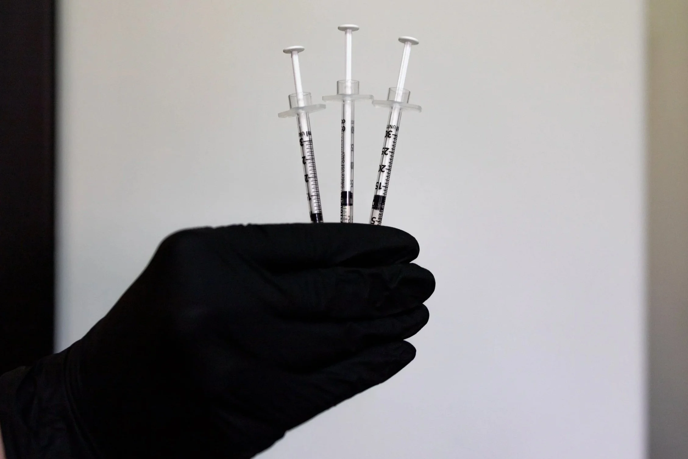 A gloved hand holding three syringes with needles directed upward, against a plain, light-colored background.