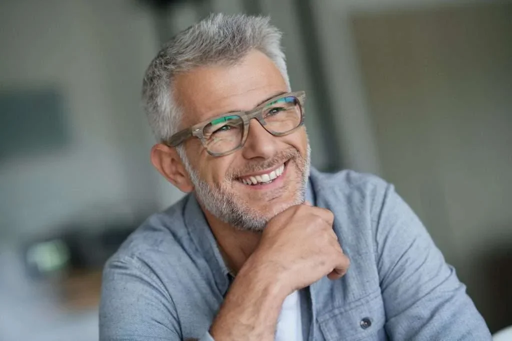 Smiling middle-aged man with gray hair and beard, wearing glasses, casual blue shirt, resting chin on hand, in a well-lit indoor setting.
