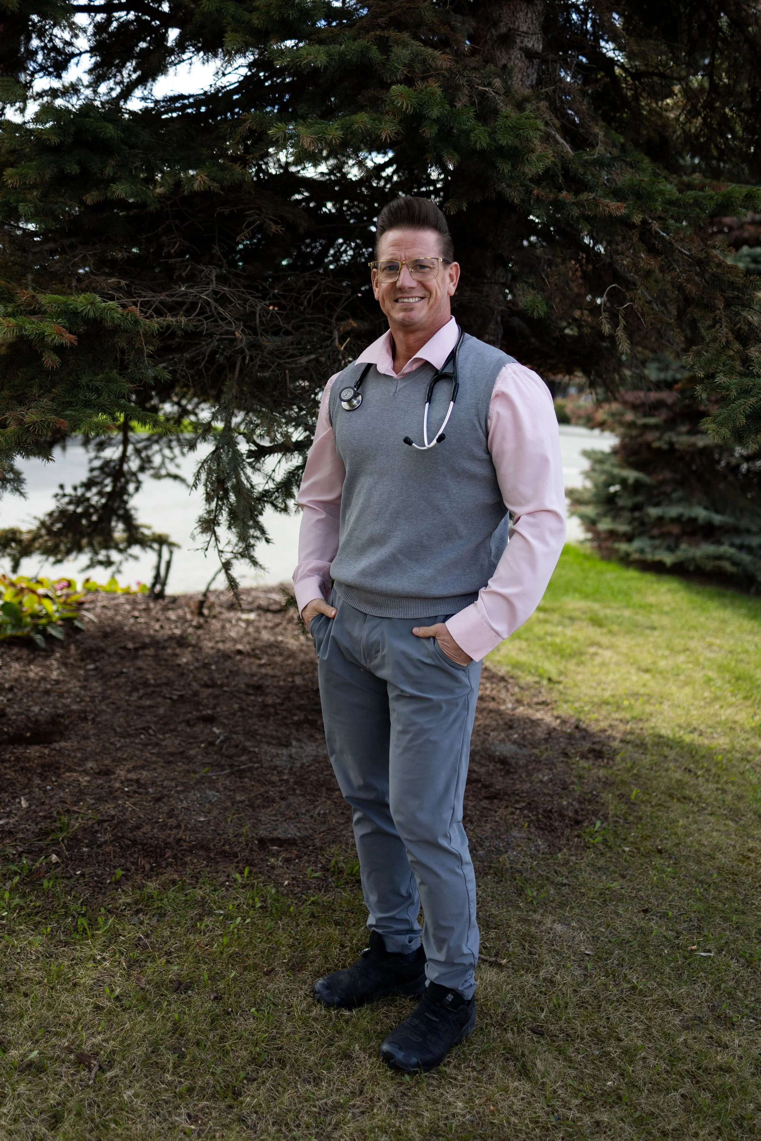 A man dressed as a doctor standing outdoors in front of a large pine tree. He is wearing glasses, a pink dress shirt, a grey vest, grey pants, and black shoes. He has a stethoscope around his neck, smiling with one hand in his pocket.