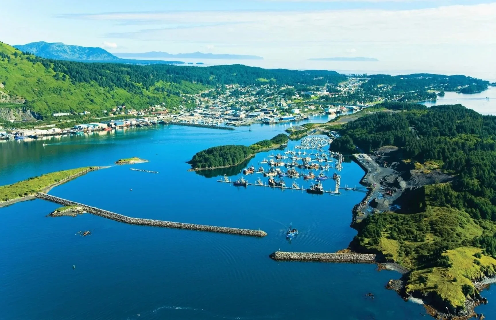Aerial view of a coastal town with a marina, surrounded by green hills and water, under a partly cloudy sky.