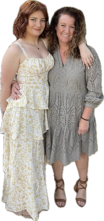 A young woman with curly red hair and an older woman with curly brown hair pose together, smiling. The young woman wears a sleeveless, yellow and white floral dress with ruffles, and the older woman wears a gray, eyelet-patterned dress with three-quarter sleeves. The older woman has her arm around the young woman's shoulder, and the young woman's hand is on her hip.