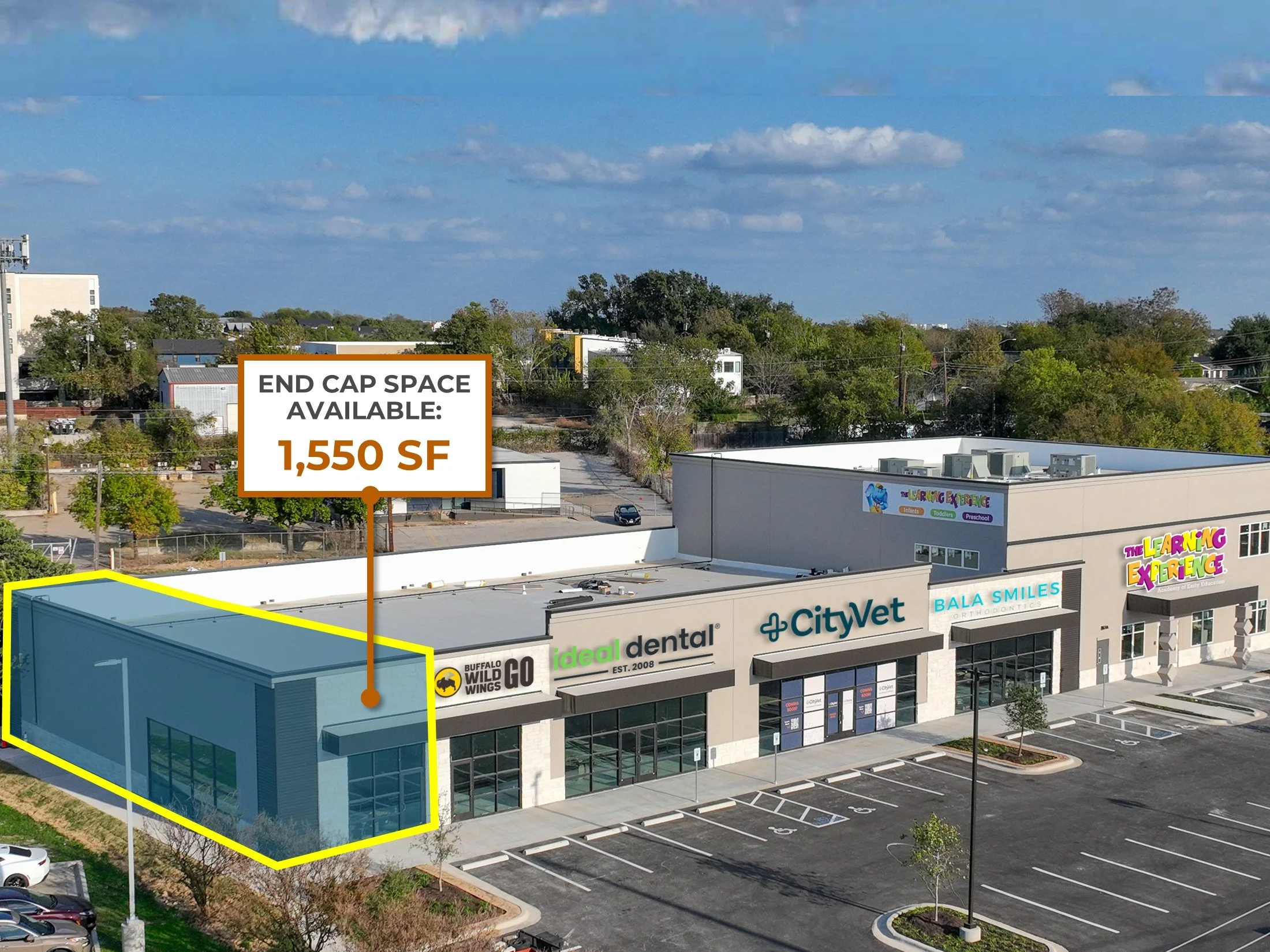 An aerial view of a commercial shopping plaza with several stores, including a CityVet, Buffalo Wild Wings, and a dental office. A large sign indicates an available space of 1,550 square feet. There are parking spaces in front of the stores and some trees and greenery surrounding the area.