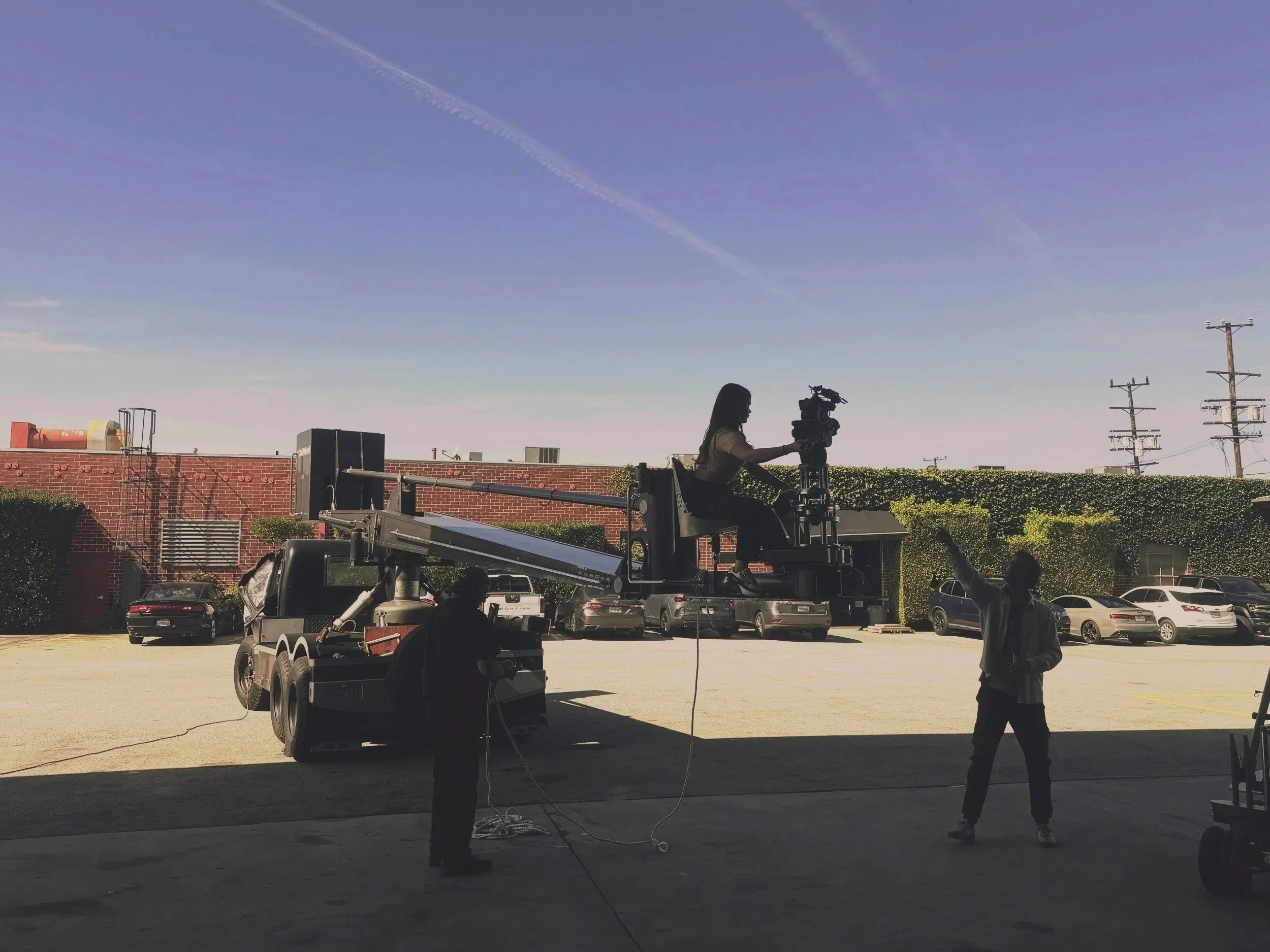 A person operating a film camera on a dolly on an outdoor set with a brick building, parked cars, and power lines in the background. A crew member is standing nearby.