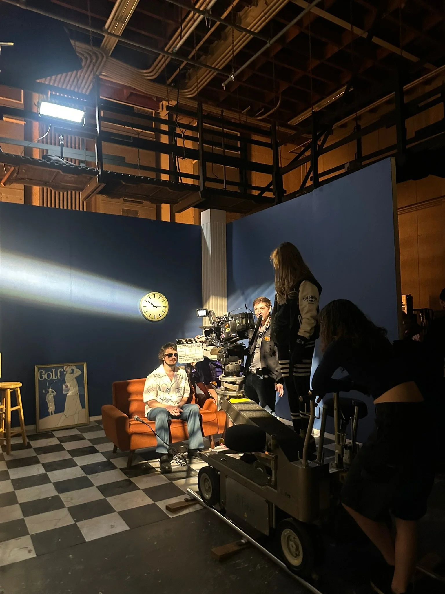 Film set with a man sitting on a brown couch, crew members and equipment surrounding him, camera, filming lights, and a blue backdrop with a clock and framed poster in a studio with checkered floor and industrial ceiling.