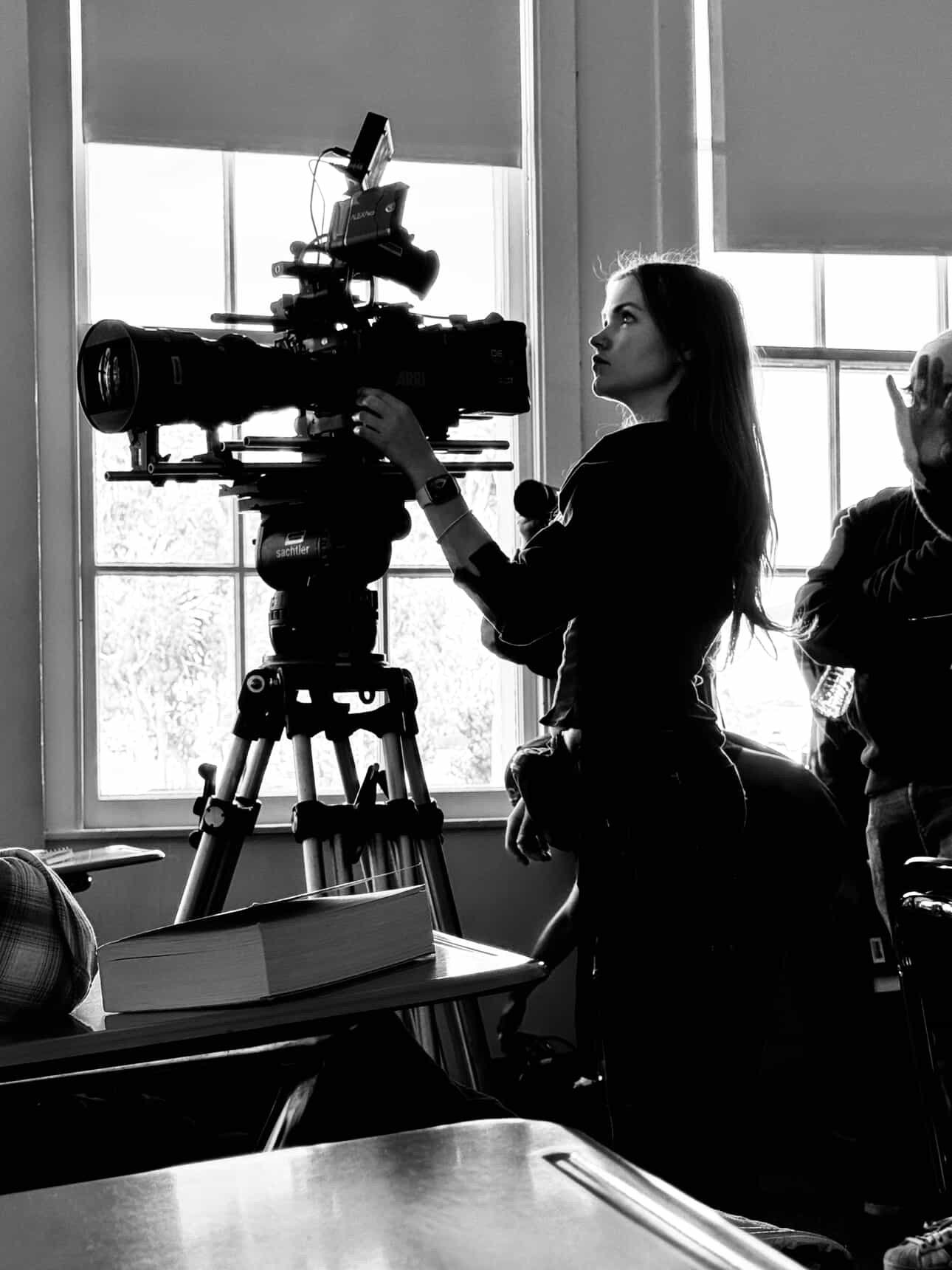 A woman with long hair operating a professional video camera on a tripod in front of a window in a classroom or meeting room.