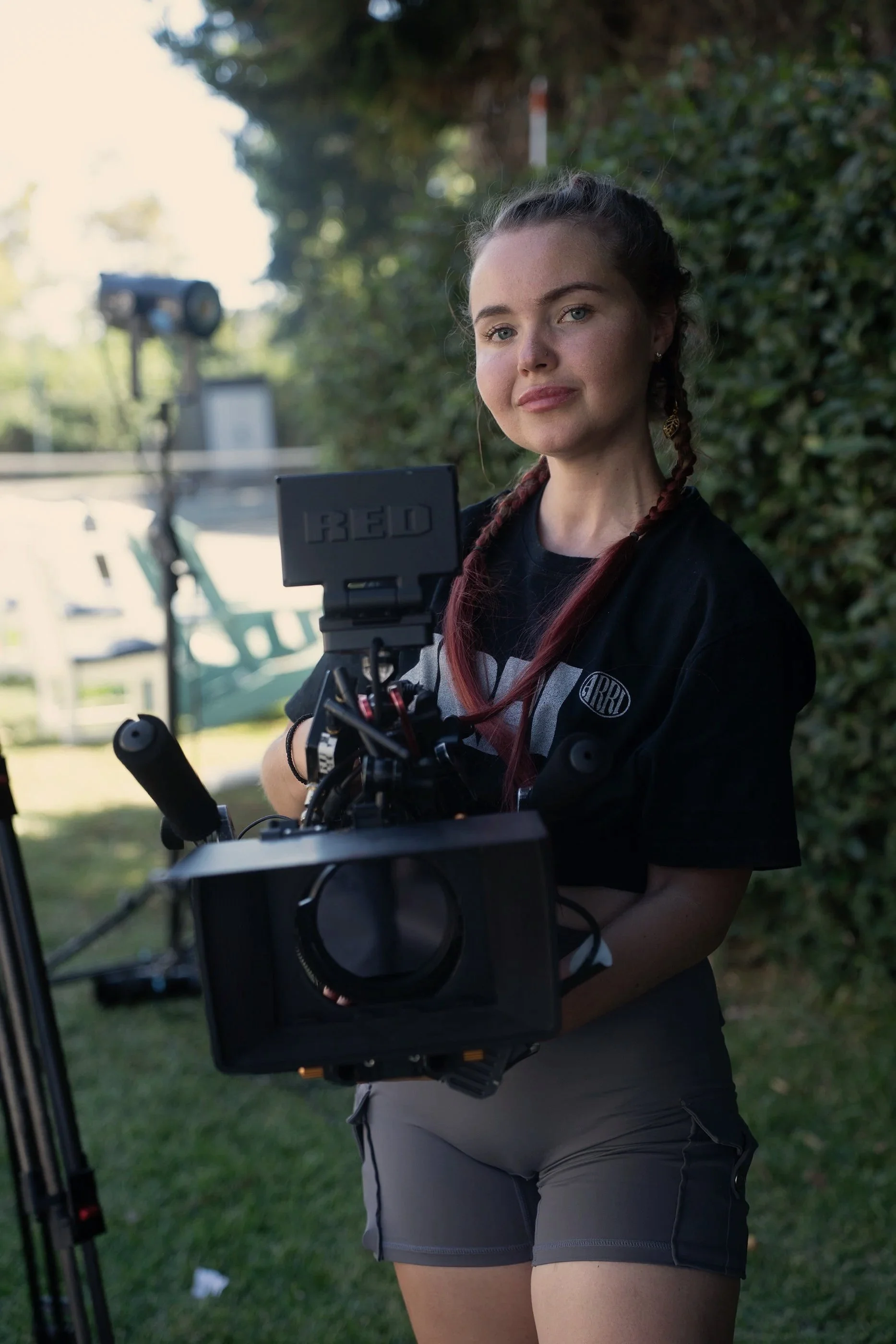 Young woman with brown hair and braided pigtails standing outdoors, holding a professional camera rig, with greenery and trees in the background.