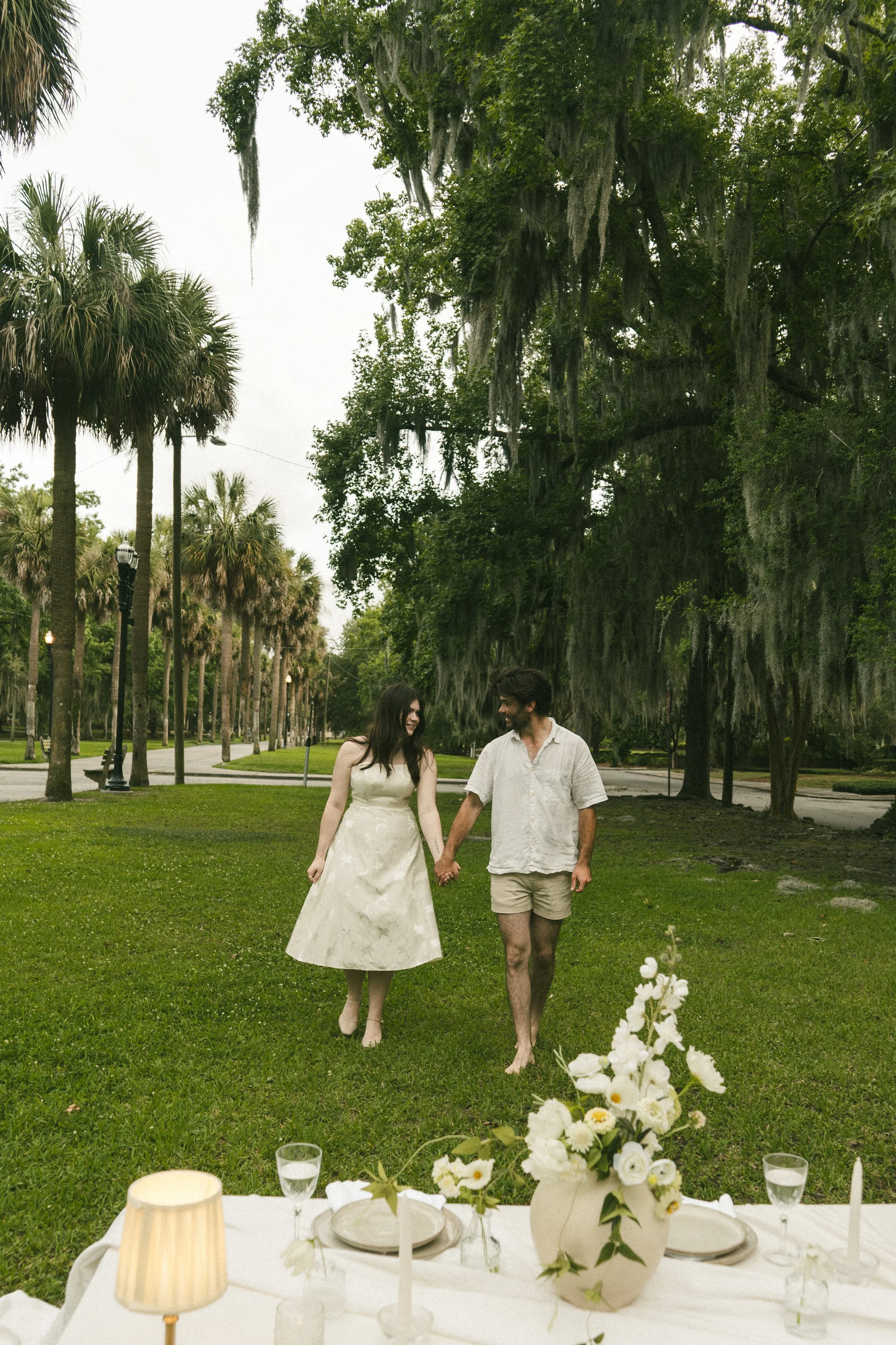 A couple holding hands walking across a lush garden with large trees, with a decorated table in the foreground and a floral centerpiece on the table.