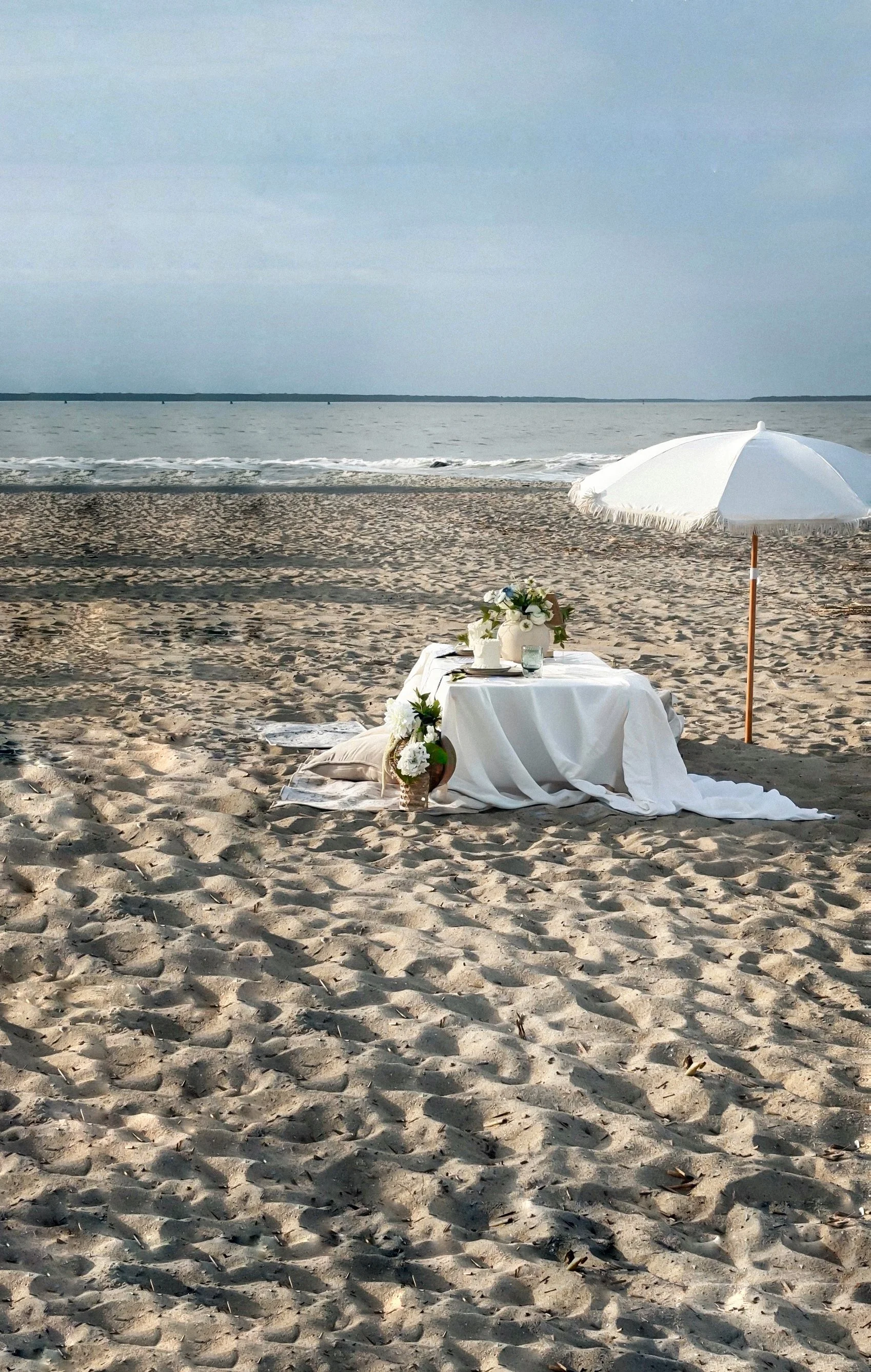 A white beach umbrella providing shade over a table decorated with white flowers, small vases, and candles on a sandy beach with the ocean in the background.