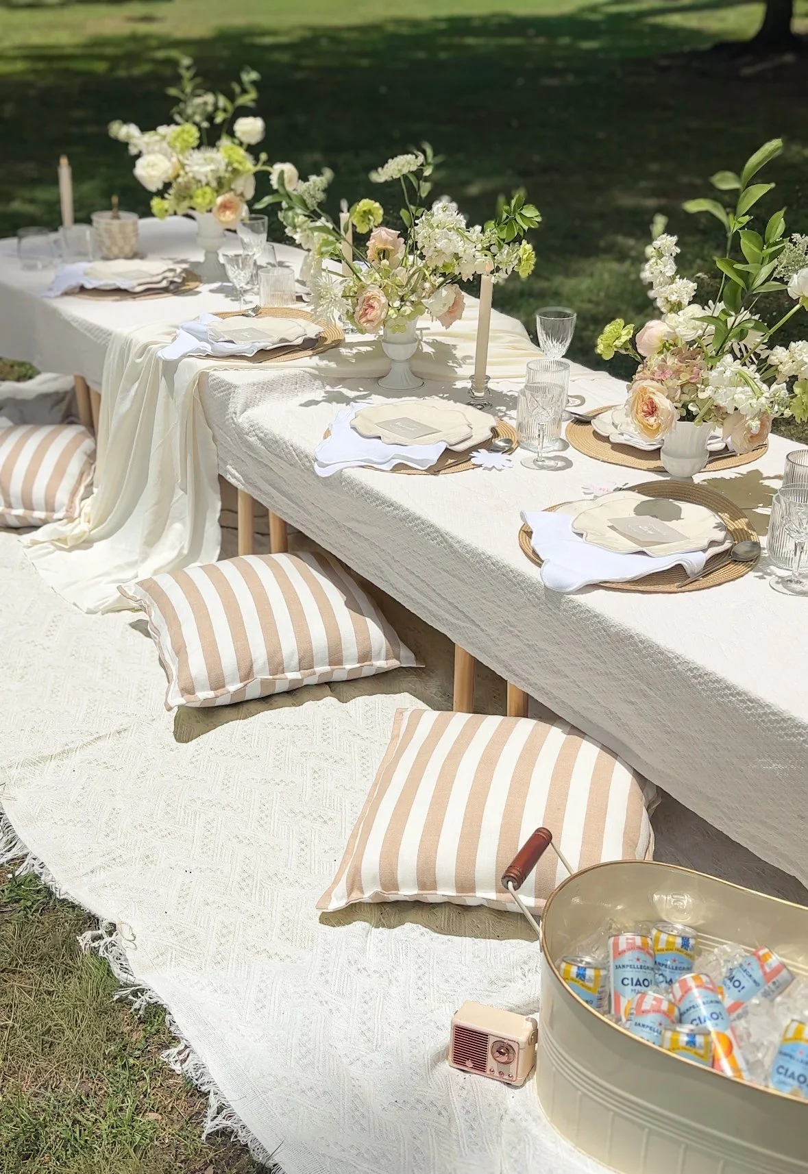 Outdoor party table decorated with white tablecloth, pink and white flowers in vases, candles, glassware, and plates with napkins, surrounded by striped cushions and pillows, with picnic items and drinks in a container nearby.