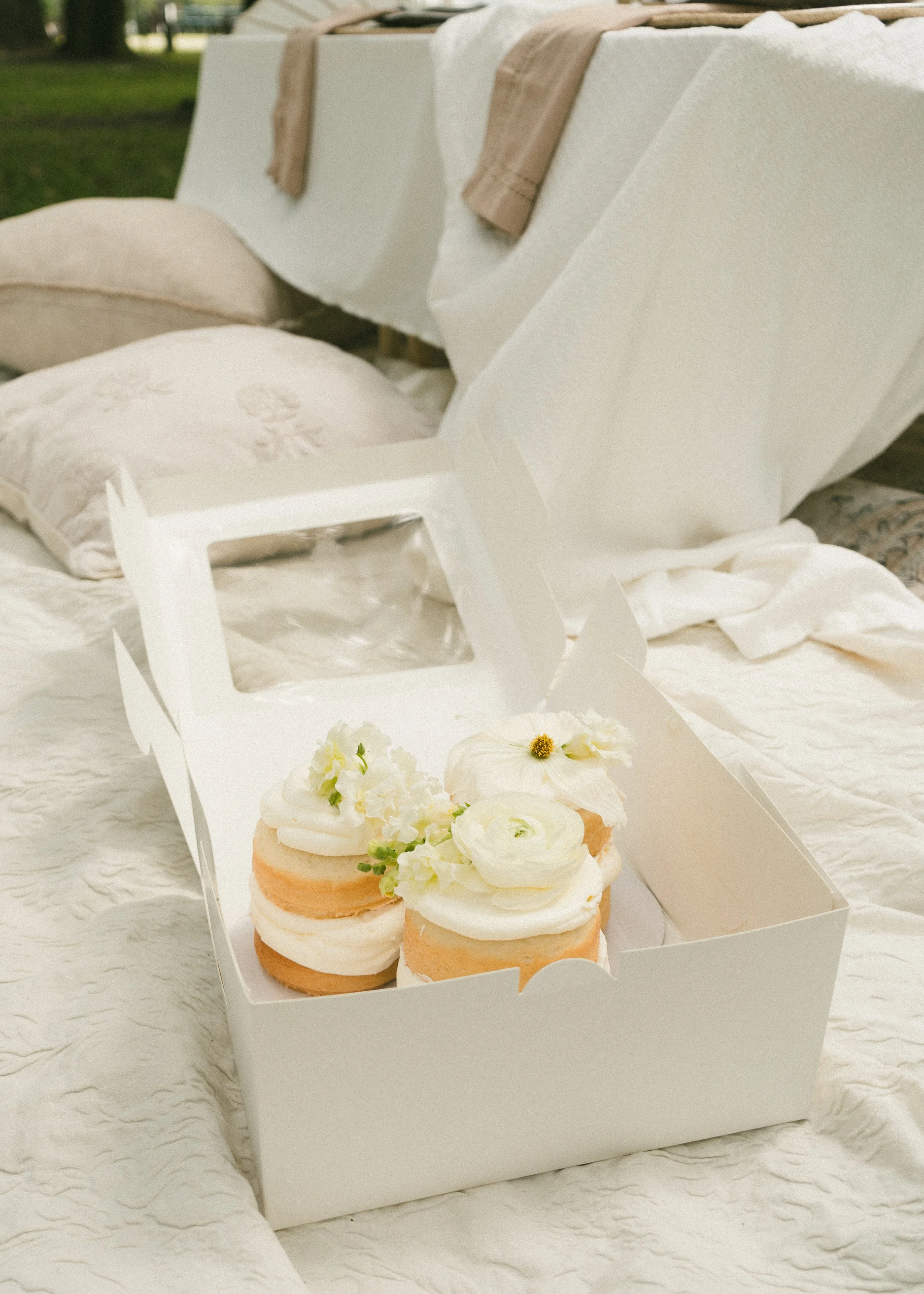 Box of vanilla cupcakes decorated with white icing and edible flowers, placed on a bed with white fabric and beige towels in the background.