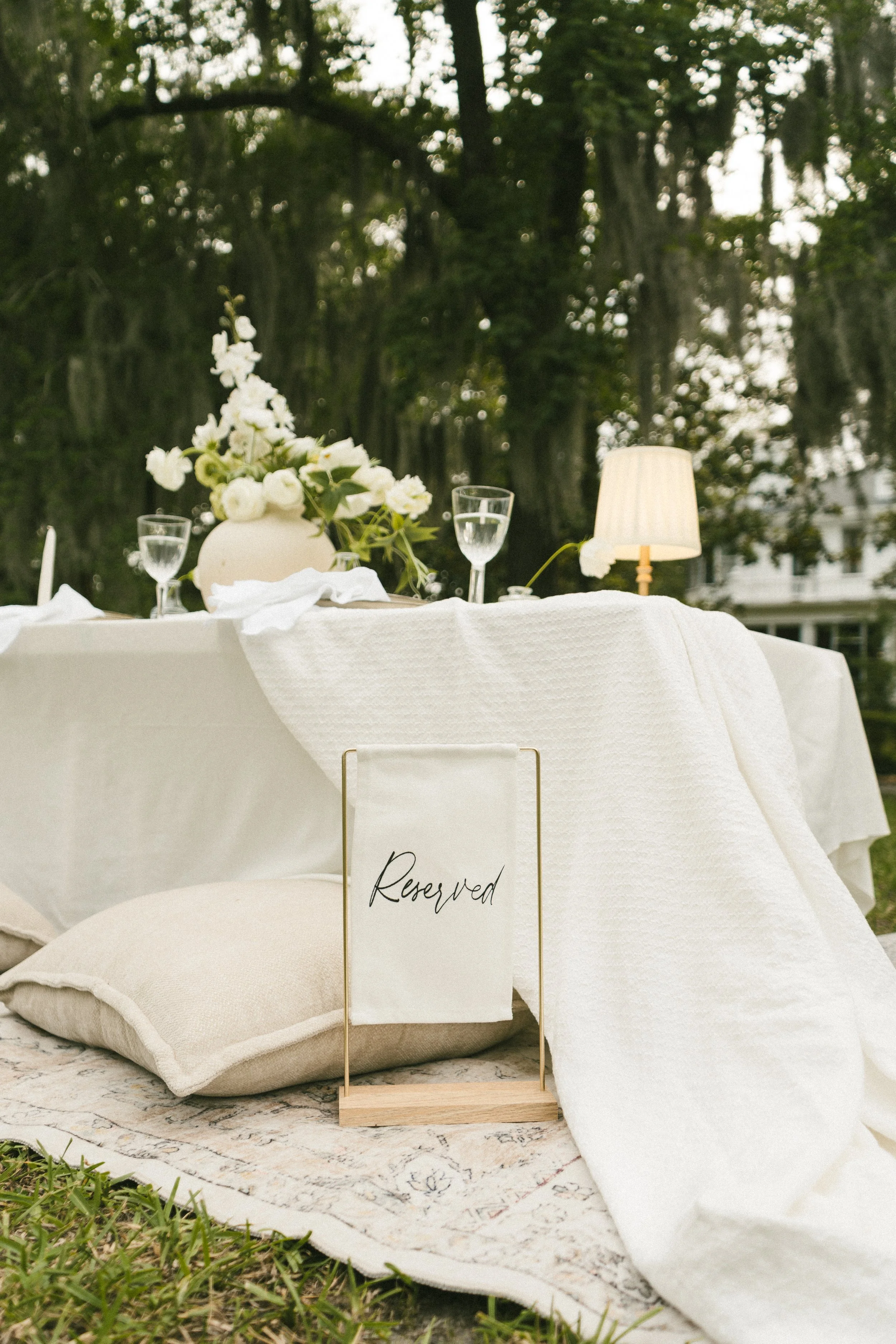 Elegant outdoor table setting with white linens, a floral centerpiece, water glasses, a lamp, and reserved sign, surrounded by greenery and trees.