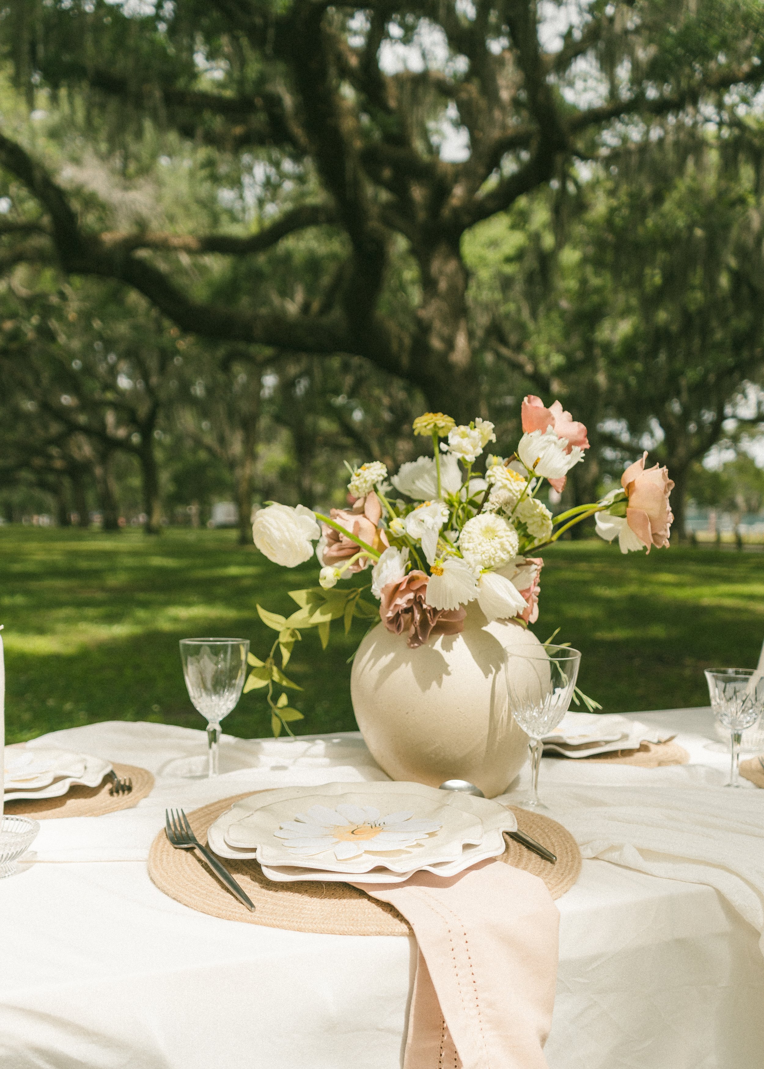 A garden party table setting with a large white vase holding a bouquet of pink, white, and cream flowers, set outdoors with trees in the background. The table has white tablecloths, woven placemats, glassware, and painted paper plates with a floral design.