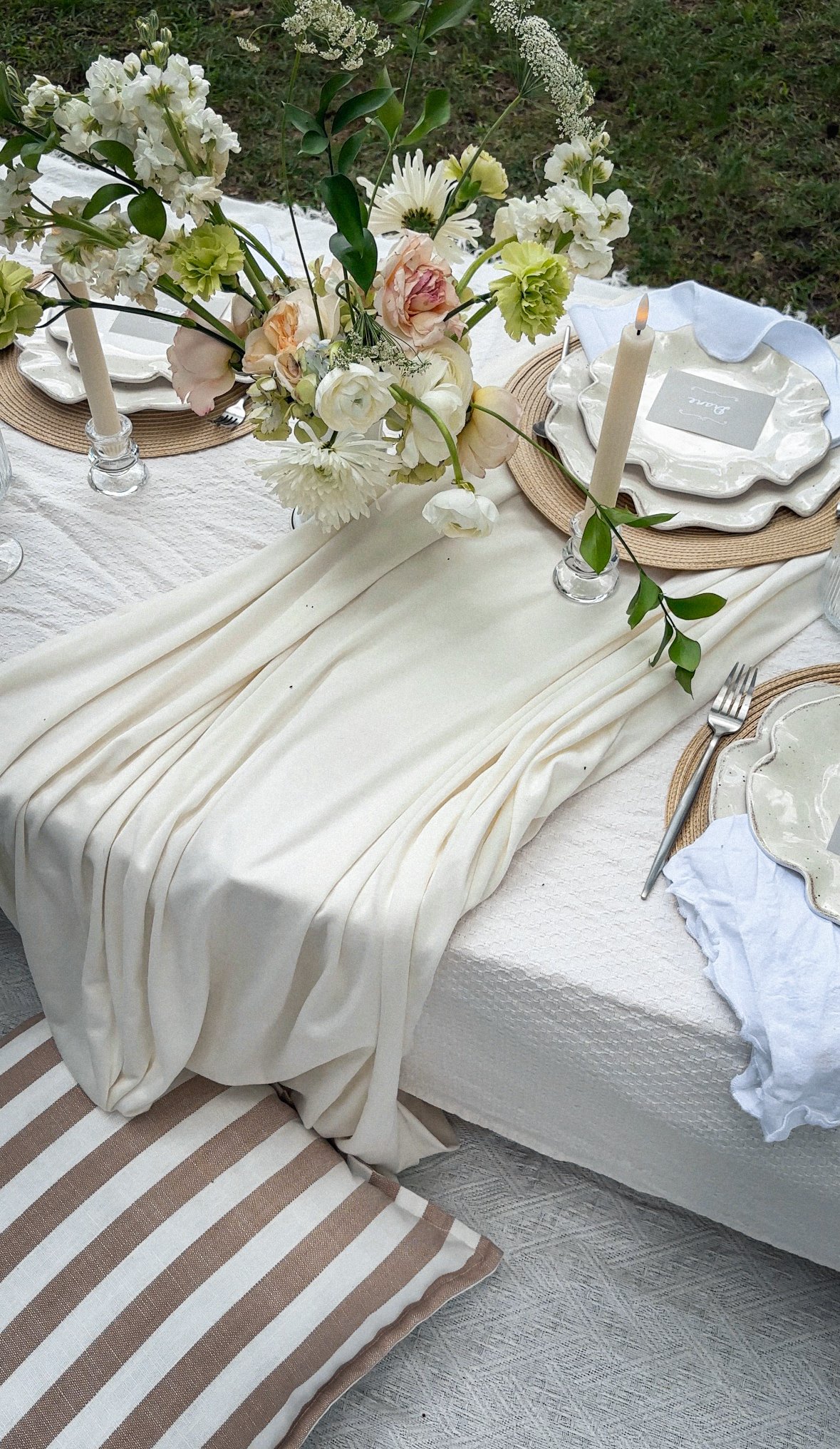 A table set for outdoor dining with a beige and white striped cushion at the end, a white tablecloth, a flowing cream-colored fabric runner, a floral centerpiece with white and pale pink flowers, candles, plates with a label reading "Love," silverware, and a white napkin, on a grassy outdoor area.
