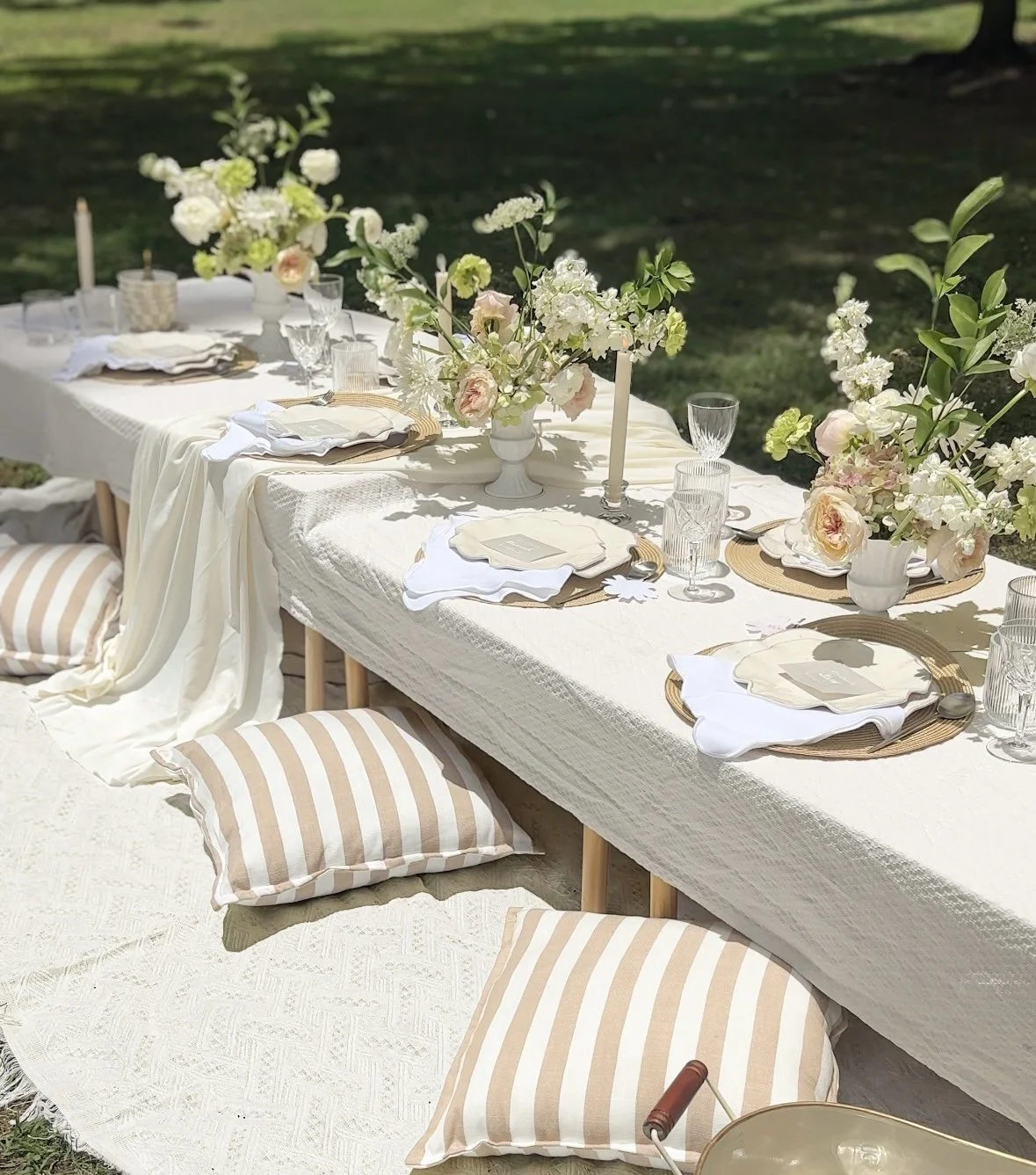 A beautifully set outdoor dining table with floral centerpieces, candles, glasses, plates, and striped cushions on the ground for seating, situated on a grassy area.