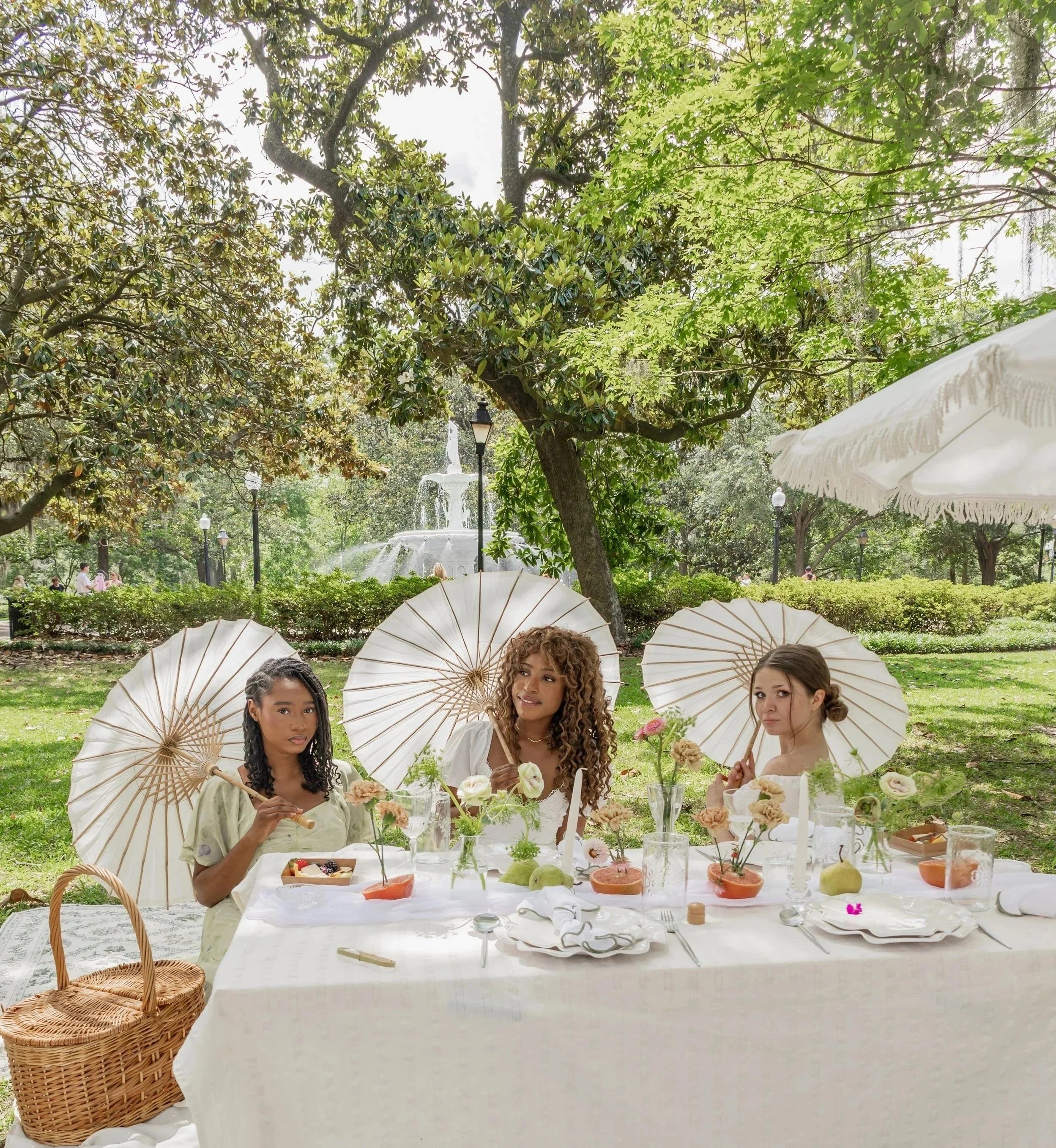 Three women sitting at a white table outdoors under trees, holding white parasols, with flowers and food on the table for a garden tea party.