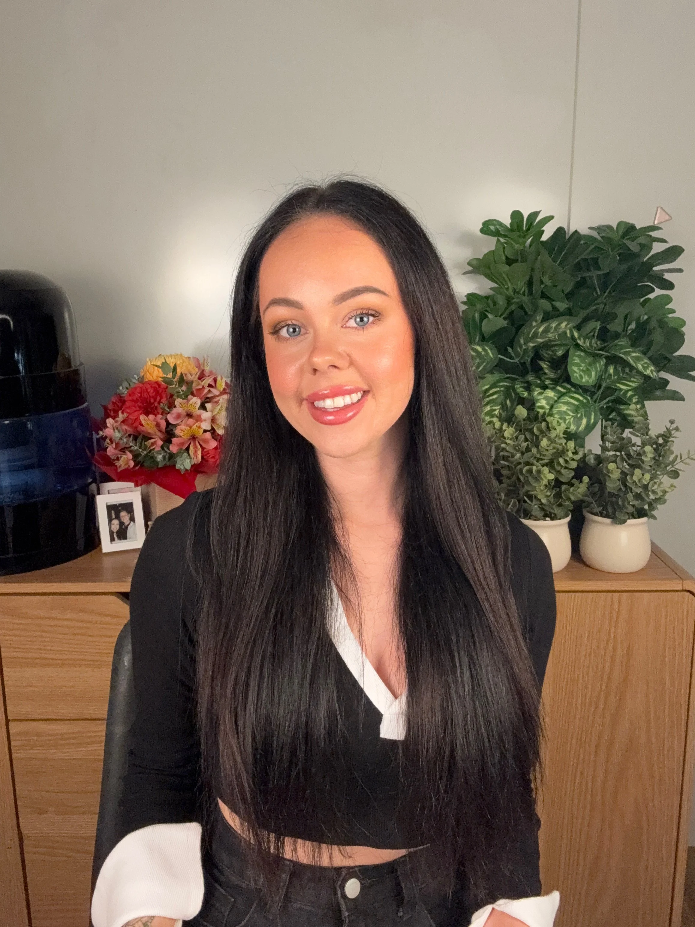 A woman with long dark hair and blue eyes smiling, sitting indoors with a wooden dresser and a large green plant in the background.