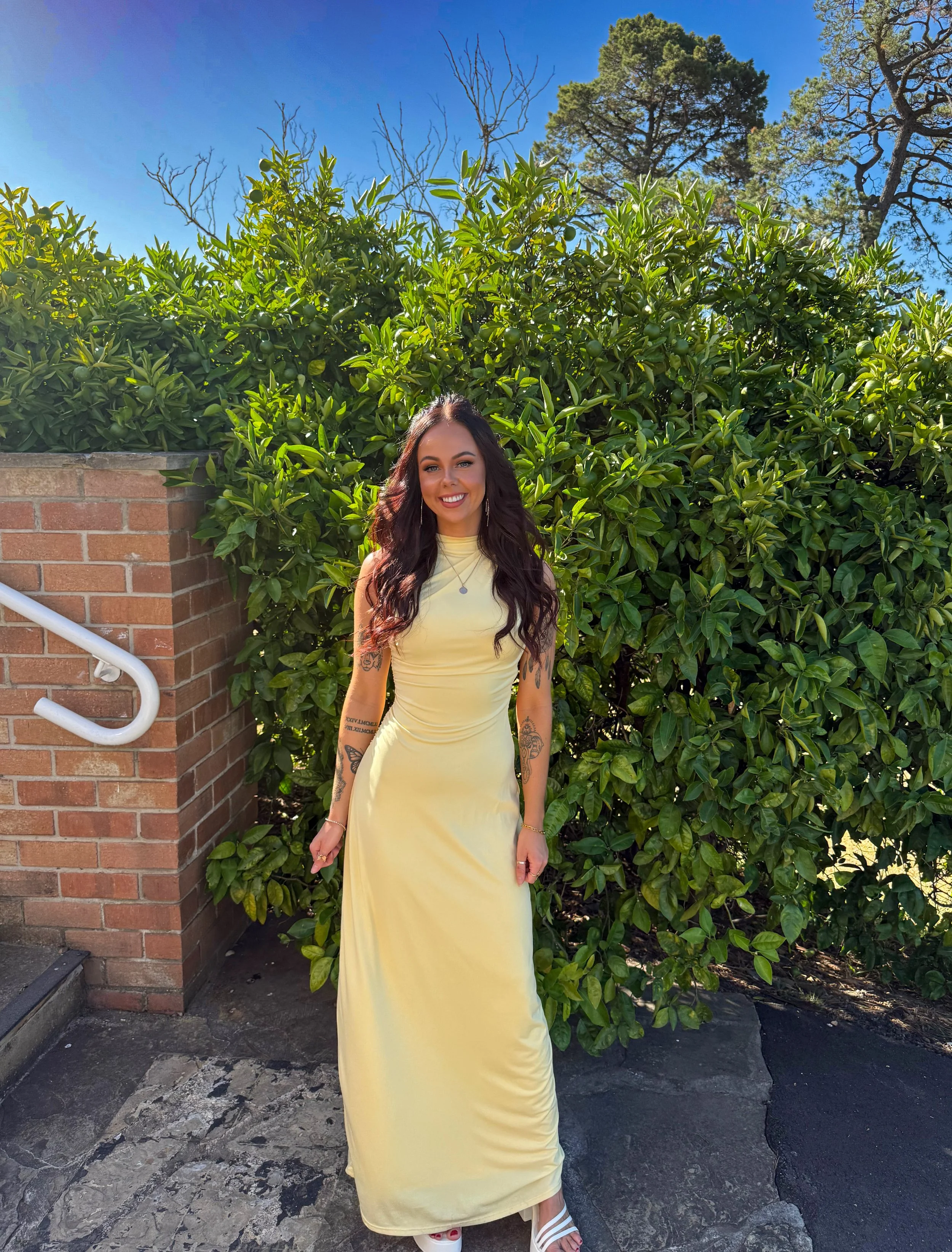 A woman with long dark wavy hair, wearing a yellow sleeveless dress and white sandals, standing outdoors in front of lush green bushes and tall trees, smiling at the camera on a sunny day.