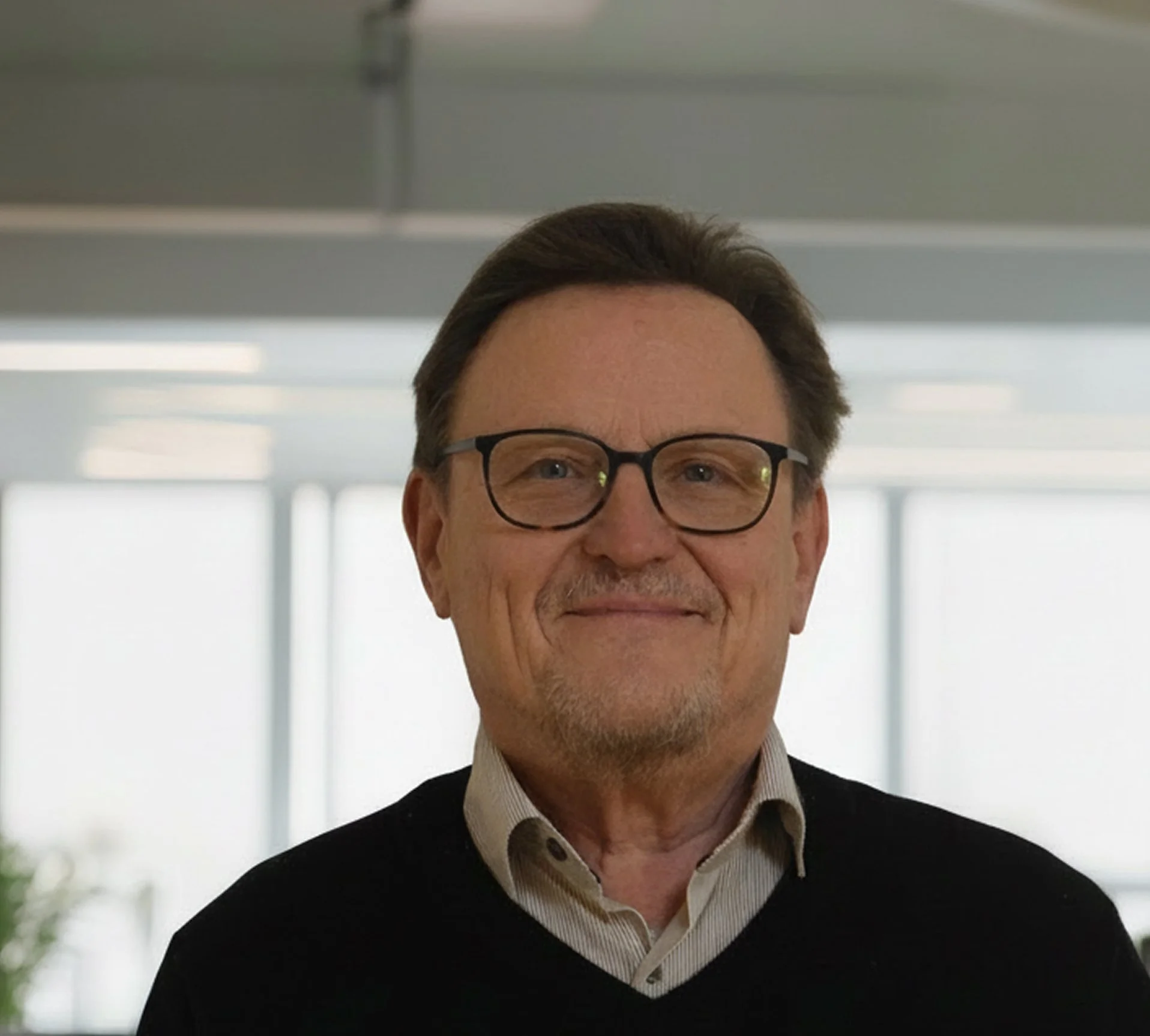 Porträt eines älteren Mannes mit Brille, dunklem Pullover und Krawatte, im Büro mit großen Fenstern im Hintergrund.