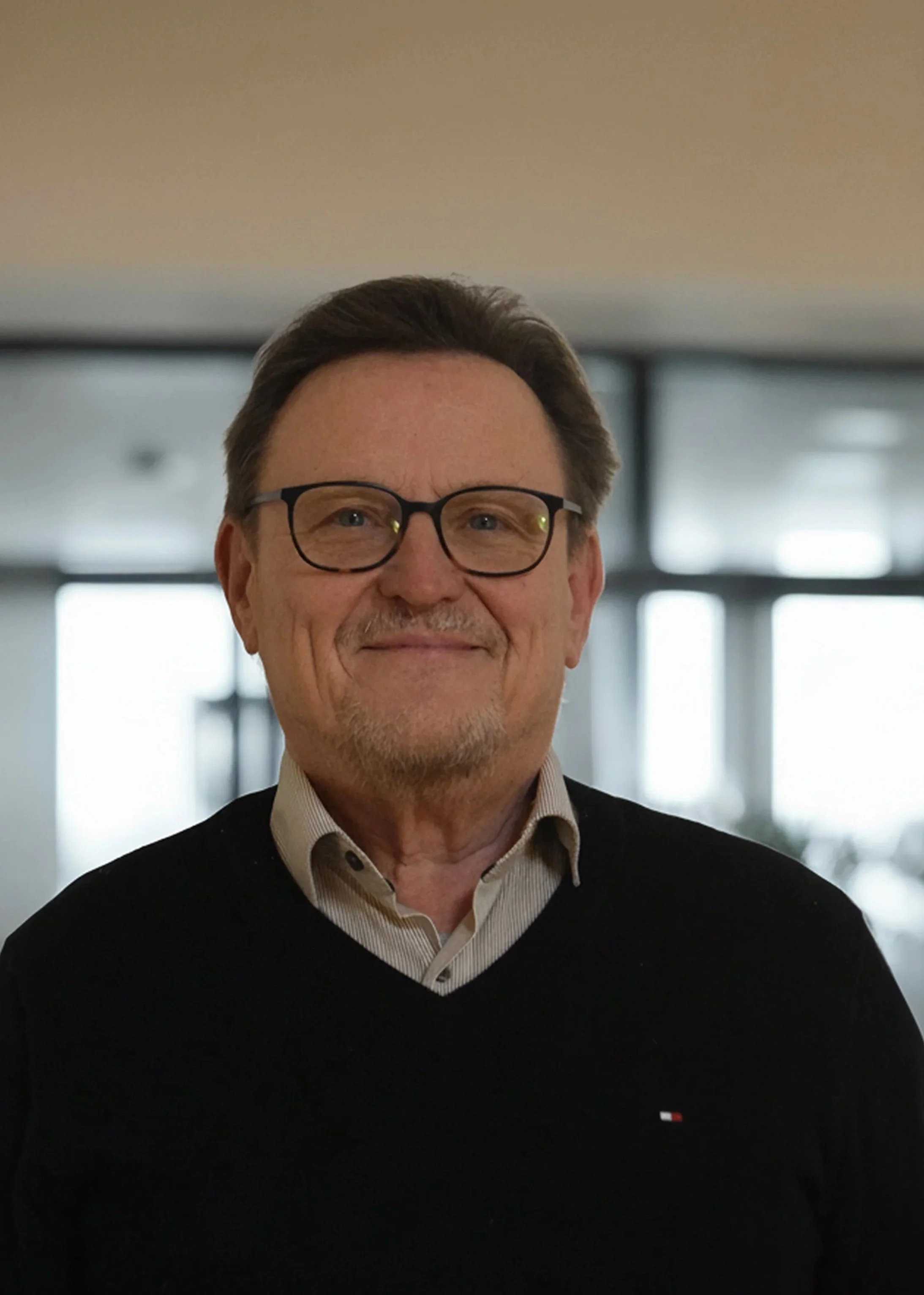 Ein älterer Mann mit braunen Haaren, Brille und einem freundlichen Ausdruck, trägt einen schwarzen Pullover mit einem kleinen Markenlogo, steht vor einer Fensterfront in einem Gebäude.