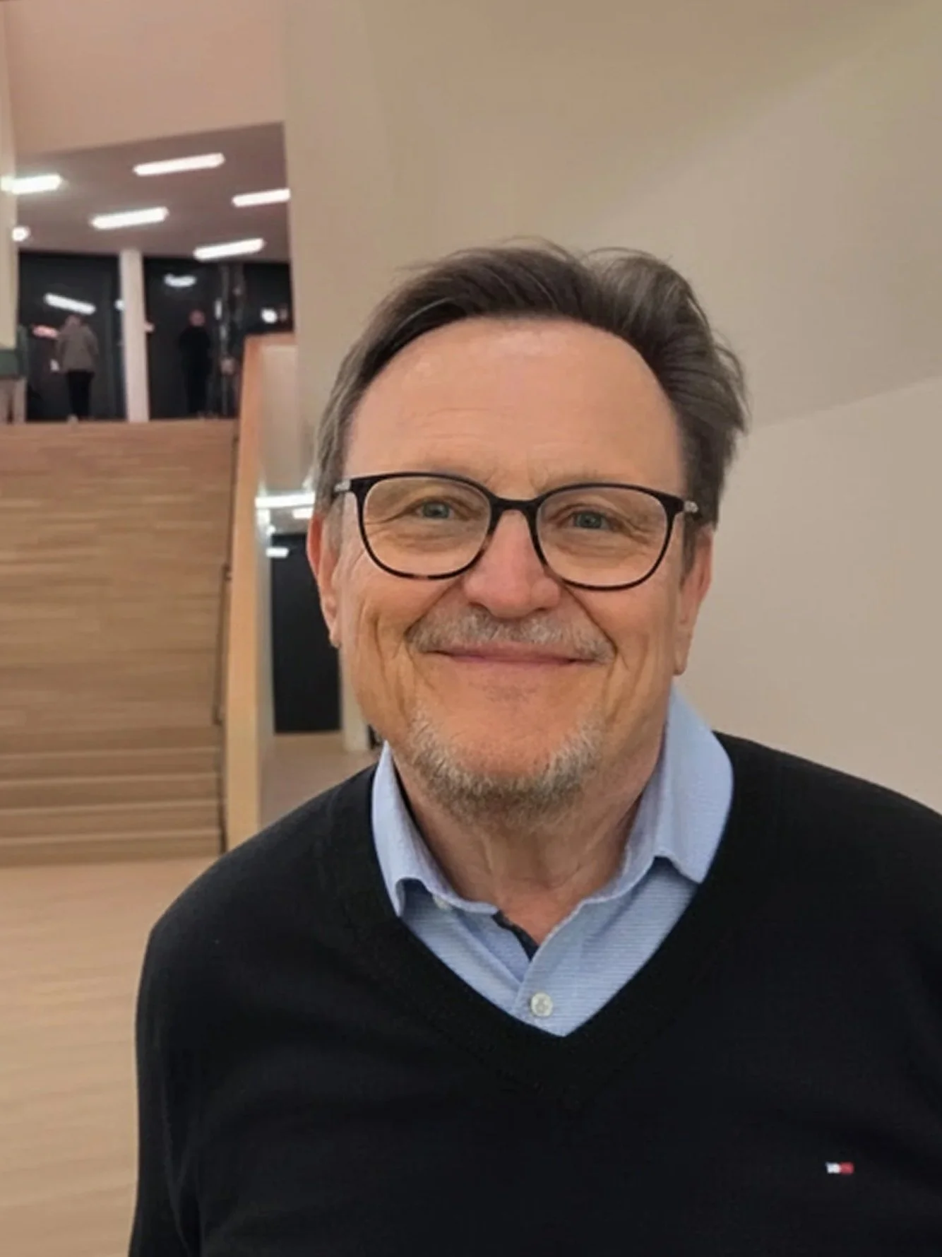 Ein lächelnder Mann mit Brille, dunklem Pullover und Hemd, sitzt in einem modernen Gebäude mit Stufen und lichtdurchflutetem Innenraum.