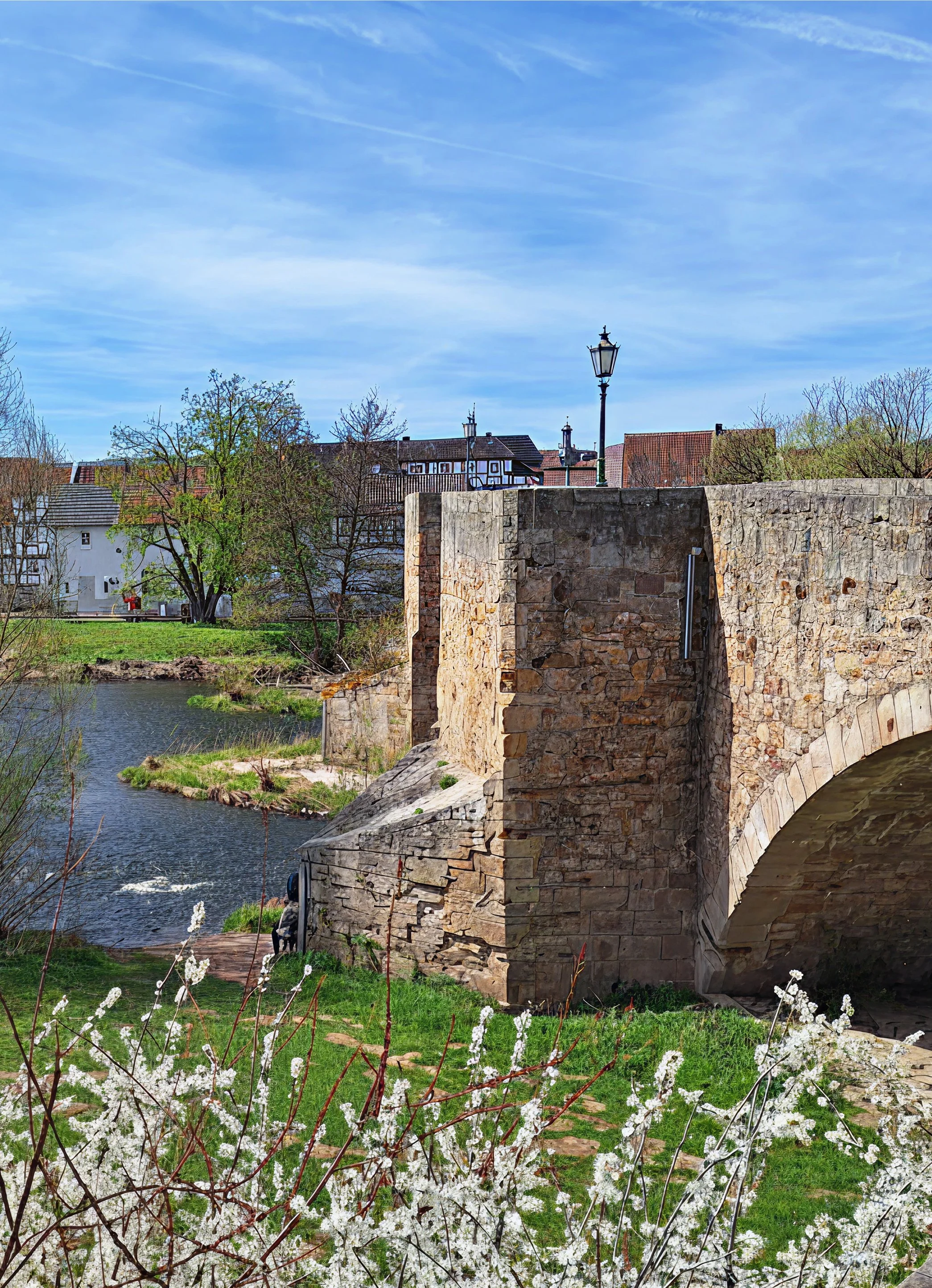 Ein Fluss fließt unter einer alten Steinbrücke hindurch. Auf der Brücke befindet sich eine Straßenlaterne. Im Hintergrund sind Hausdächer und Bäume sichtbar, einige Bäume tragen noch keine Blätter. Im Vordergrund befinden sich weiße Blütensträucher.