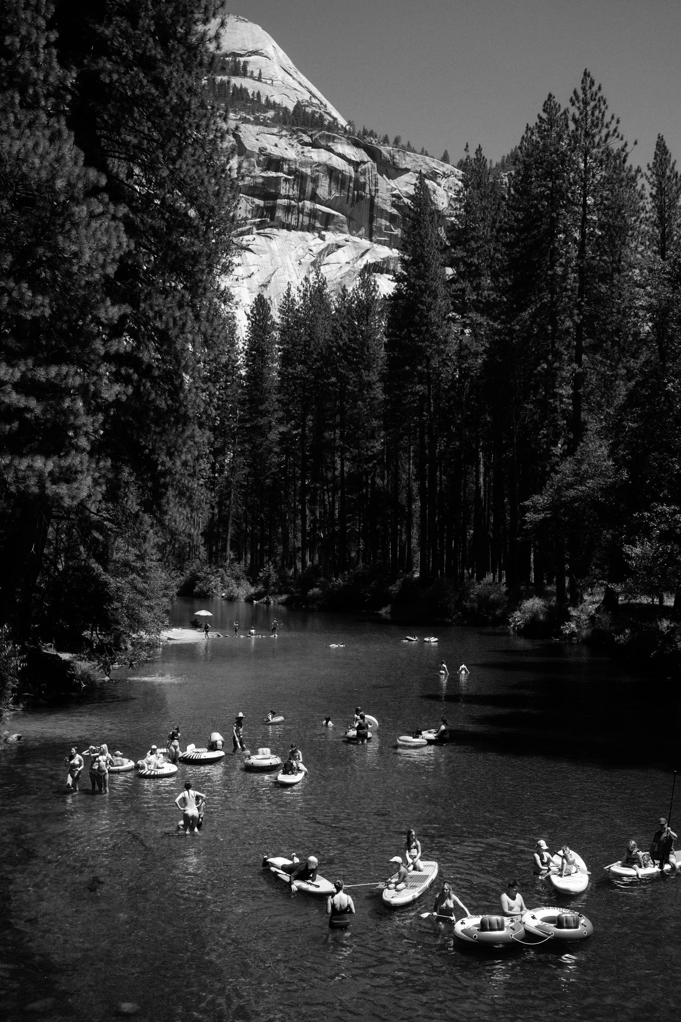 People swimming and floating on inflatable tubes and rafts in a river, surrounded by trees with a mountain in the background.