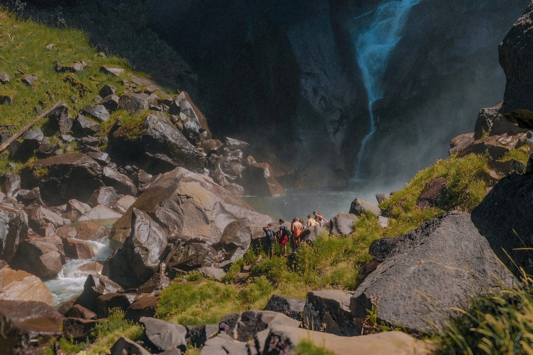 A group of people gathers on rocks near the base of a large waterfall flowing down a deep canyon with steep rocky walls, surrounded by green vegetation.