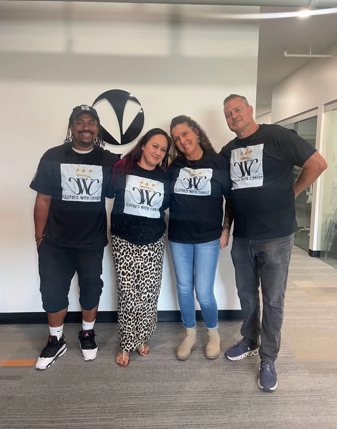 Four people wearing matching black T-shirts with a logo and the text "Clothed with Christ," standing in an office space with a white wall and company logo behind them.