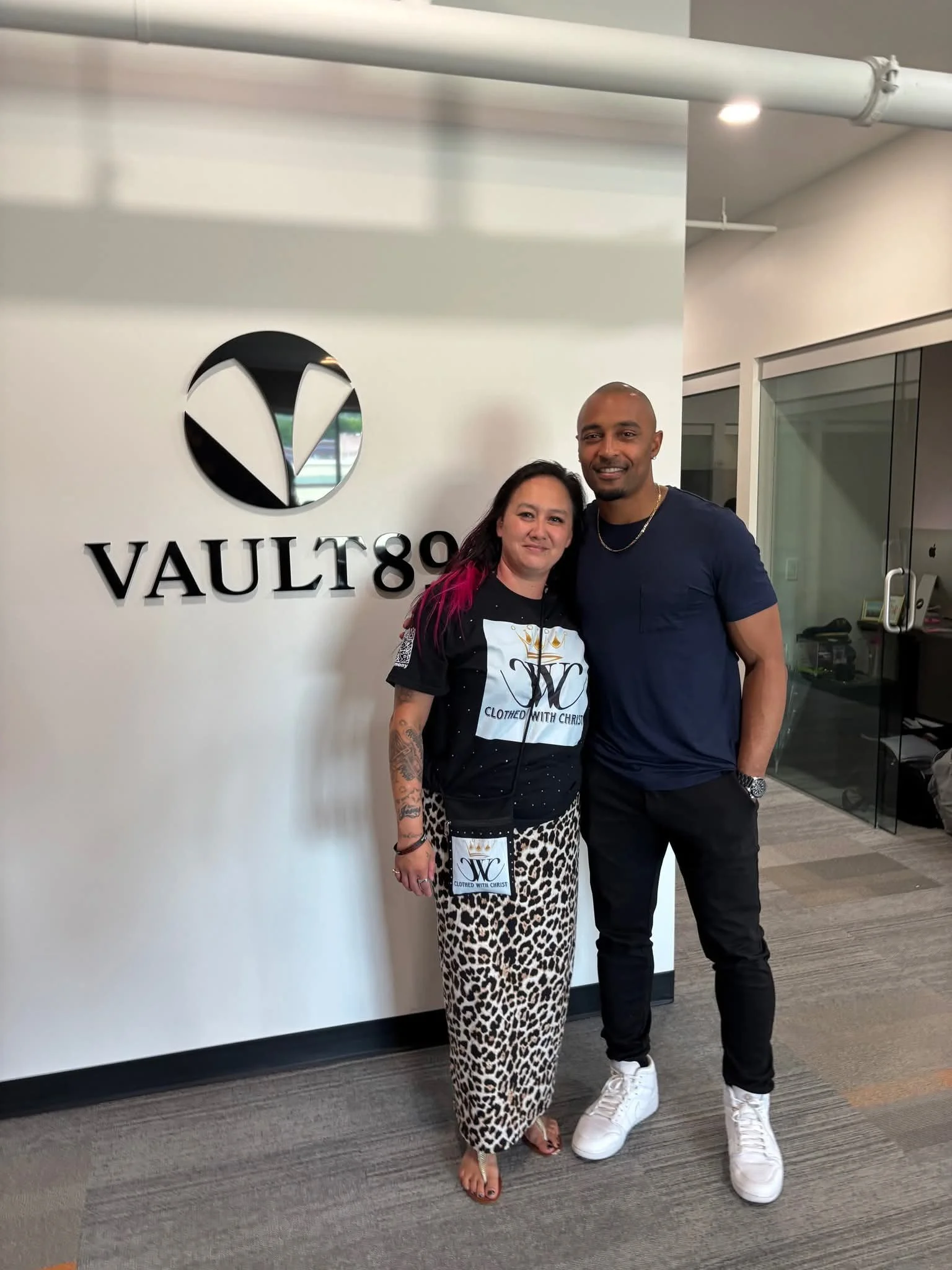 Two people standing together in front of a wall with the VAULT89 logo, smiling at the camera. The woman has dark hair with pink streaks, tattoos on her arm, and is wearing a black and white patterned skirt and shirt. The man has a shaved head, dark beard, and is wearing a navy blue t-shirt, black pants, and white sneakers.