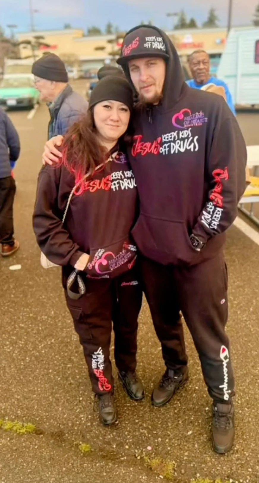 Two people wearing matching hoodies with anti-drug messages, standing outdoors at a gathering, with other people and buildings in the background.