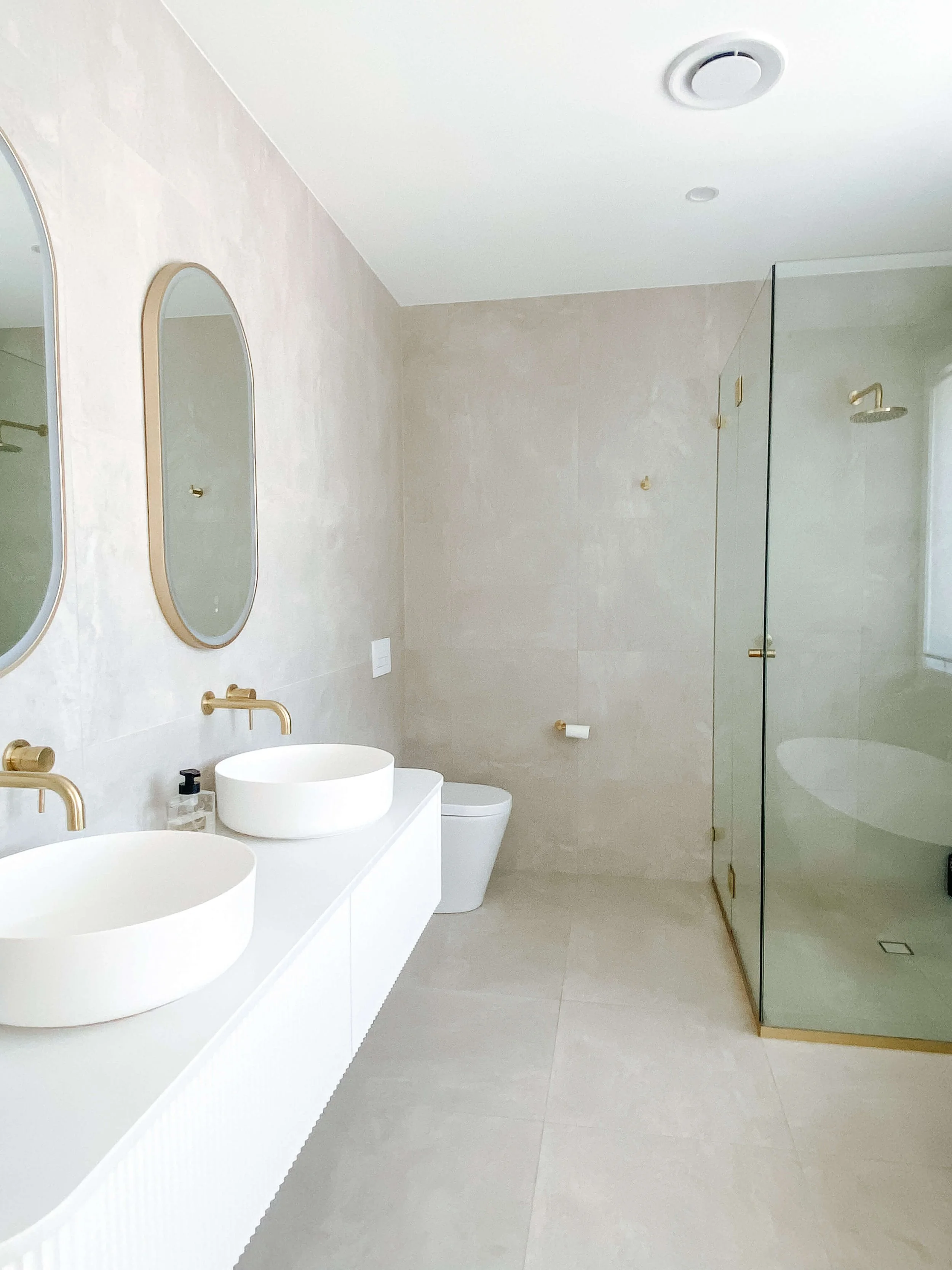 A modern bathroom with two white vessel sinks, gold fixtures, mirrors, beige tiled walls, a toilet, and a glass shower enclosure.