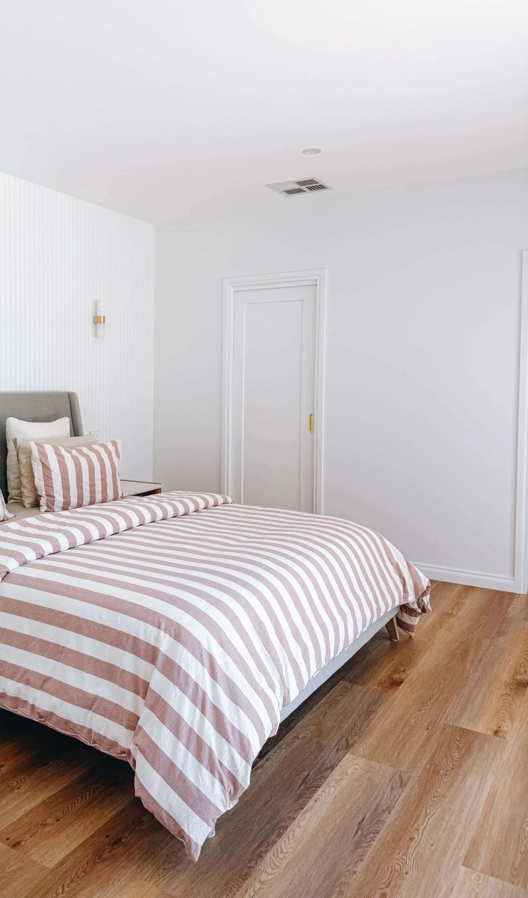 A bedroom with a bed that has striped pink and white bedding and pillows, a wooden floor, a white wall with vertical textured design, a door, and minimal decor.