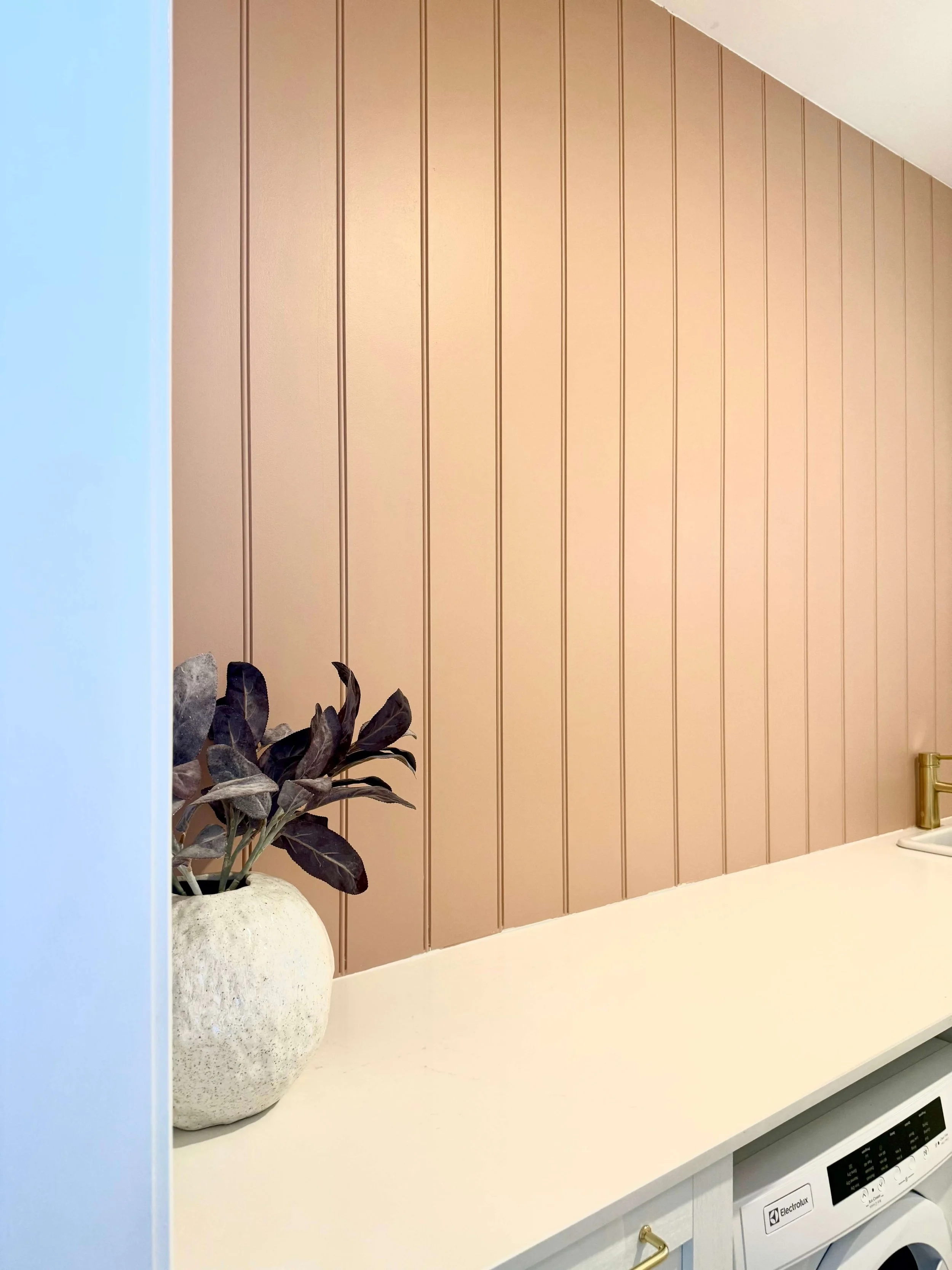 A kitchen countertop with a white potted plant with dark purple leaves, a beige textured wall paneling backdrop, a corner of a yellow-gold faucet, and a washing machine beneath the countertop.
