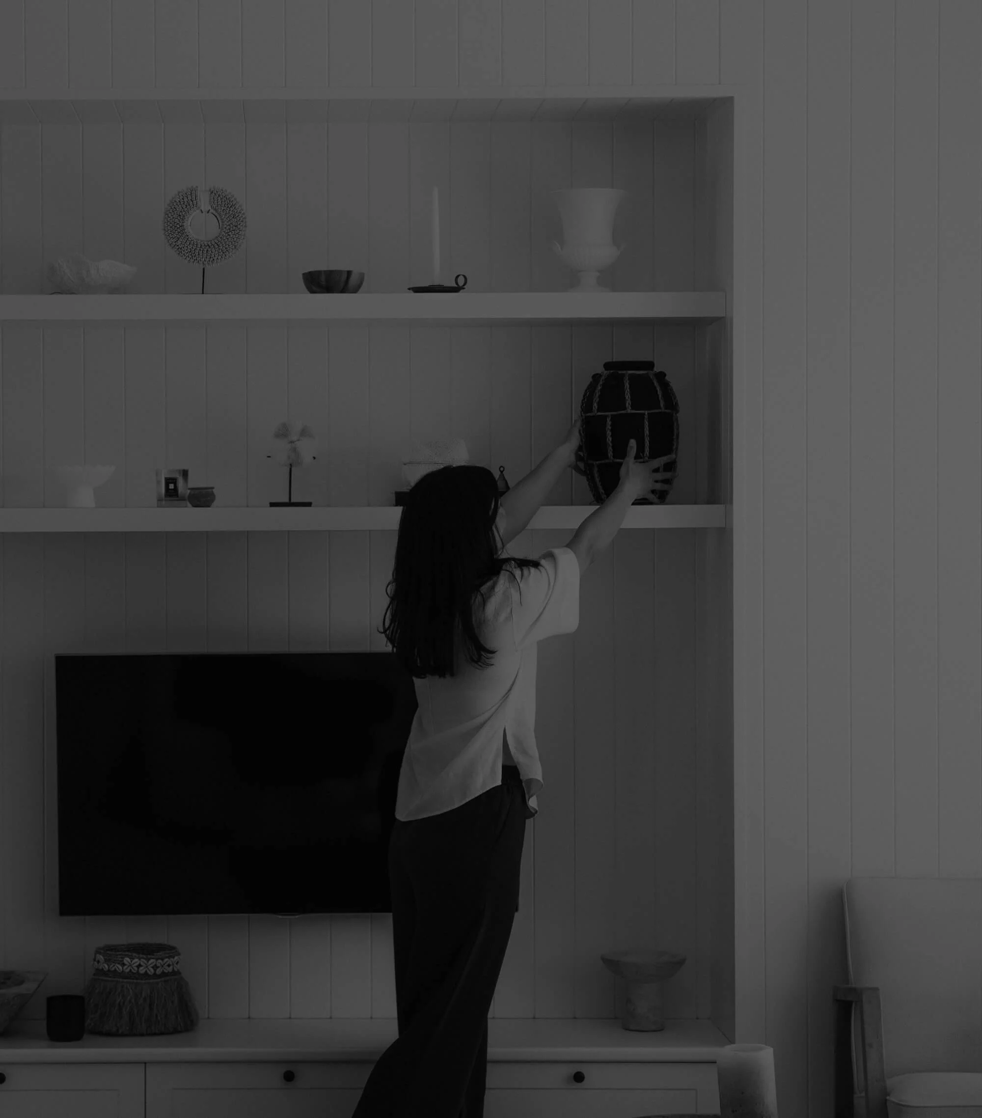A person with long dark hair reaching up towards a large decorative vase on a wooden shelf in a minimalist, white room with vertical paneling.