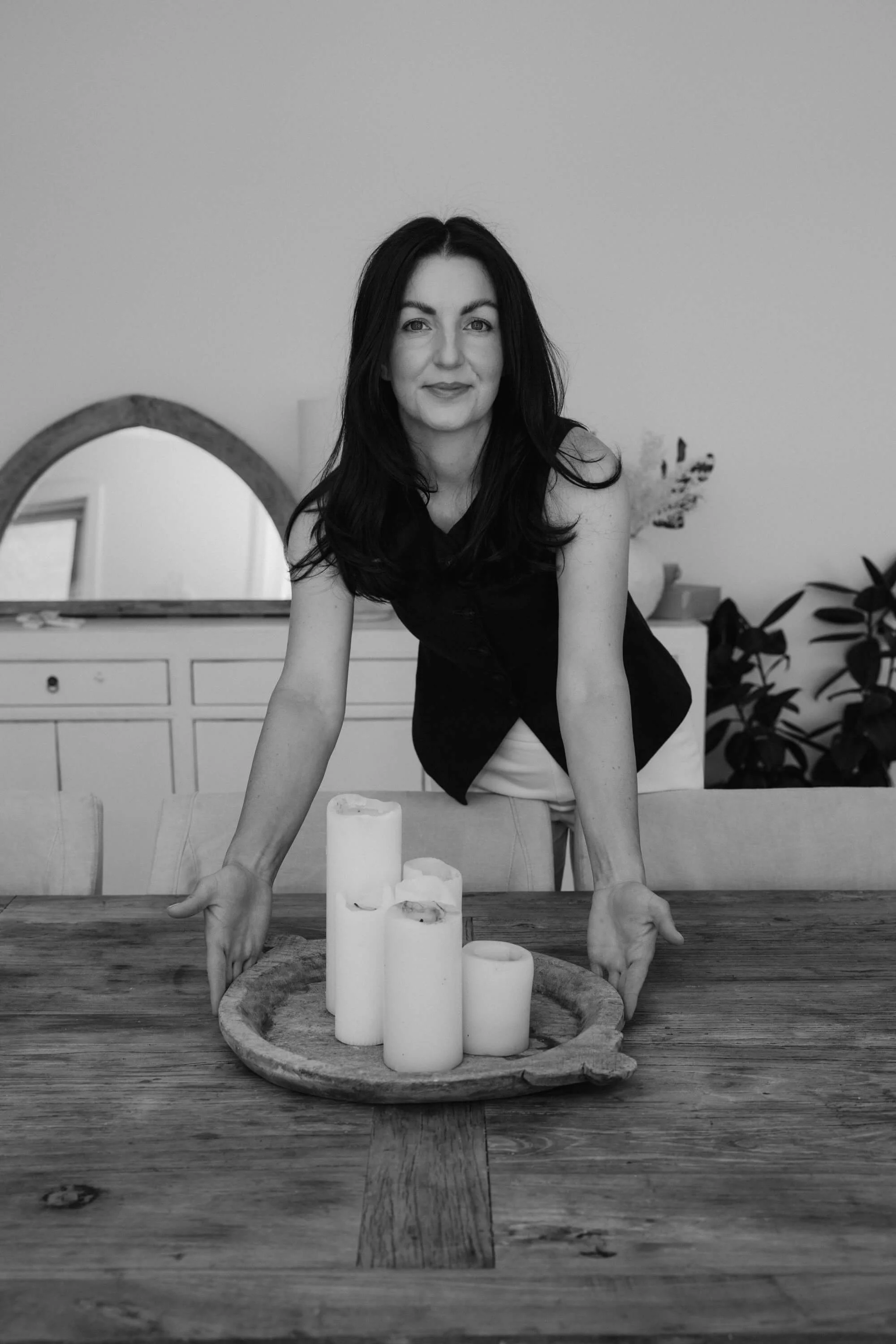 A woman with dark hair and a sleeveless top leans over a table with her hands on the surface. In front of her, there is a round wooden tray with four white candles of varying heights. The background includes a mirror, a white dresser, and some houseplants.