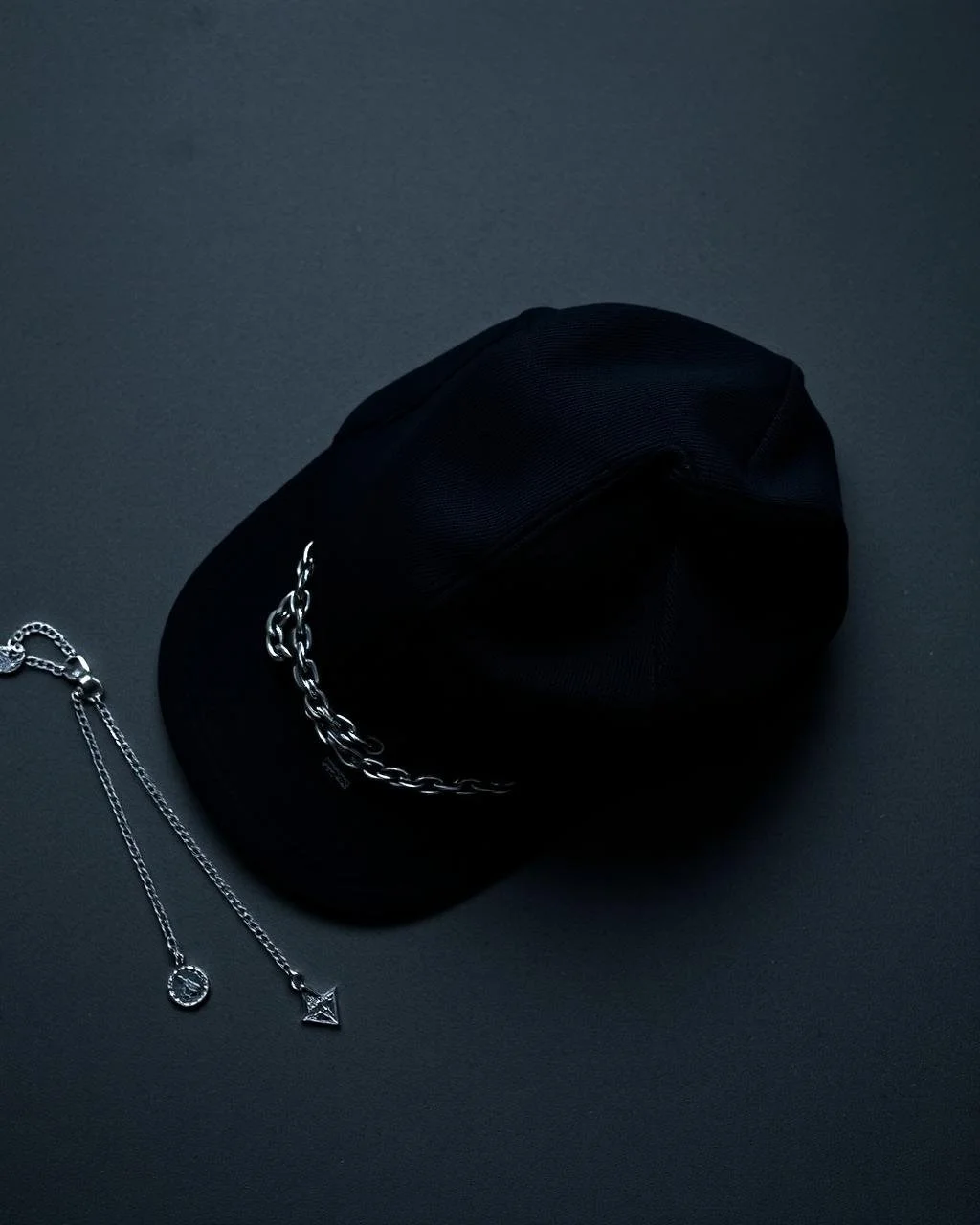 Black baseball cap with a silver chain and two silver necklaces on a dark surface.