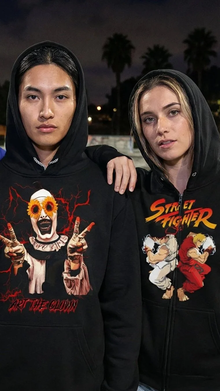Two young people wearing black hoodies with graphic designs, standing outdoors at night with palm trees in the background.