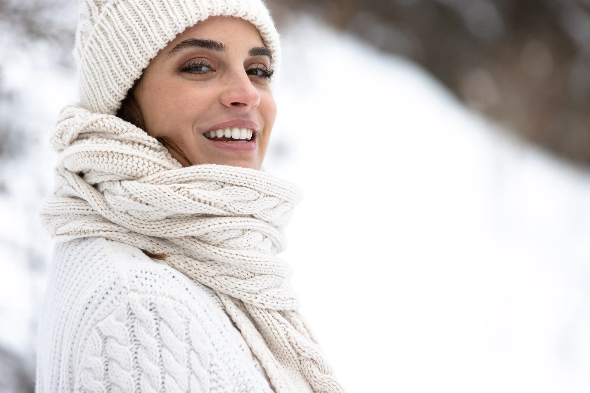 Ein lächelnder Frau trägt eine Wollmütze, einen Schal und einen Pullover, steht draussen im Winter, im Hintergrund schneebedecktes Gelände.